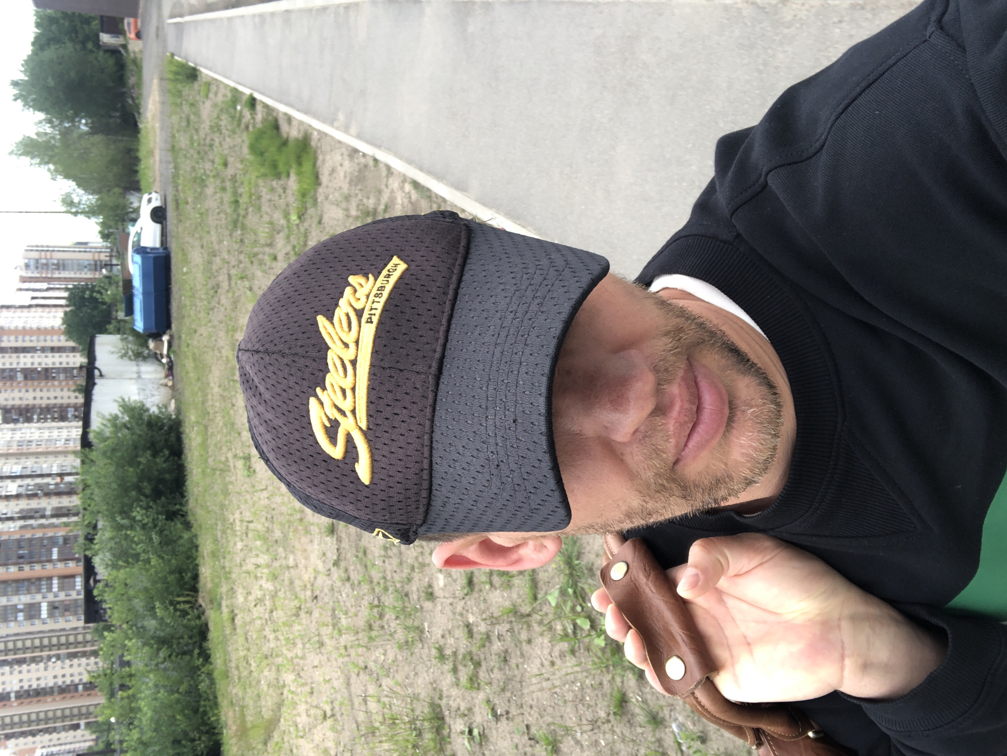 A man with a beard wearing a black baseball cap with yellow lettering and a black sweatshirt is standing outdoors. He is holding a brown leather strap with brass studs over his left shoulder. The background consists of tall apartment buildings, some trees, and a paved walkway. A blue cylindrical object and a white car are visible in the middle ground. This scene is consistent with the outskirts of Saint Petersburg, Russia, likely in a residential development area.