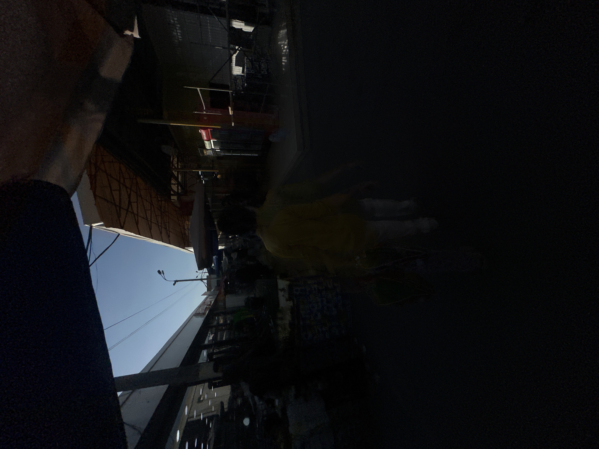 The image shows an outdoor scene, possibly a market, with people present. The orientation of the image is rotated 90 degrees counter-clockwise. In the foreground, a dark blue fabric is visible. Behind this, a structure with a patterned canopy is seen, suggesting a marketplace. Further in the background, there are buildings and what appears to be a street with a lamppost and overhead wires. The sky is clear and blue, indicating daytime. A person in a yellow garment is partially visible amidst the structures, appearing to be walking or standing. The scene is somewhat dark in certain areas, possibly due to shadows or the lighting conditions. There are some faint signs with text, but they are not legible. The location is identified as Tashkent, Uzbekistan.