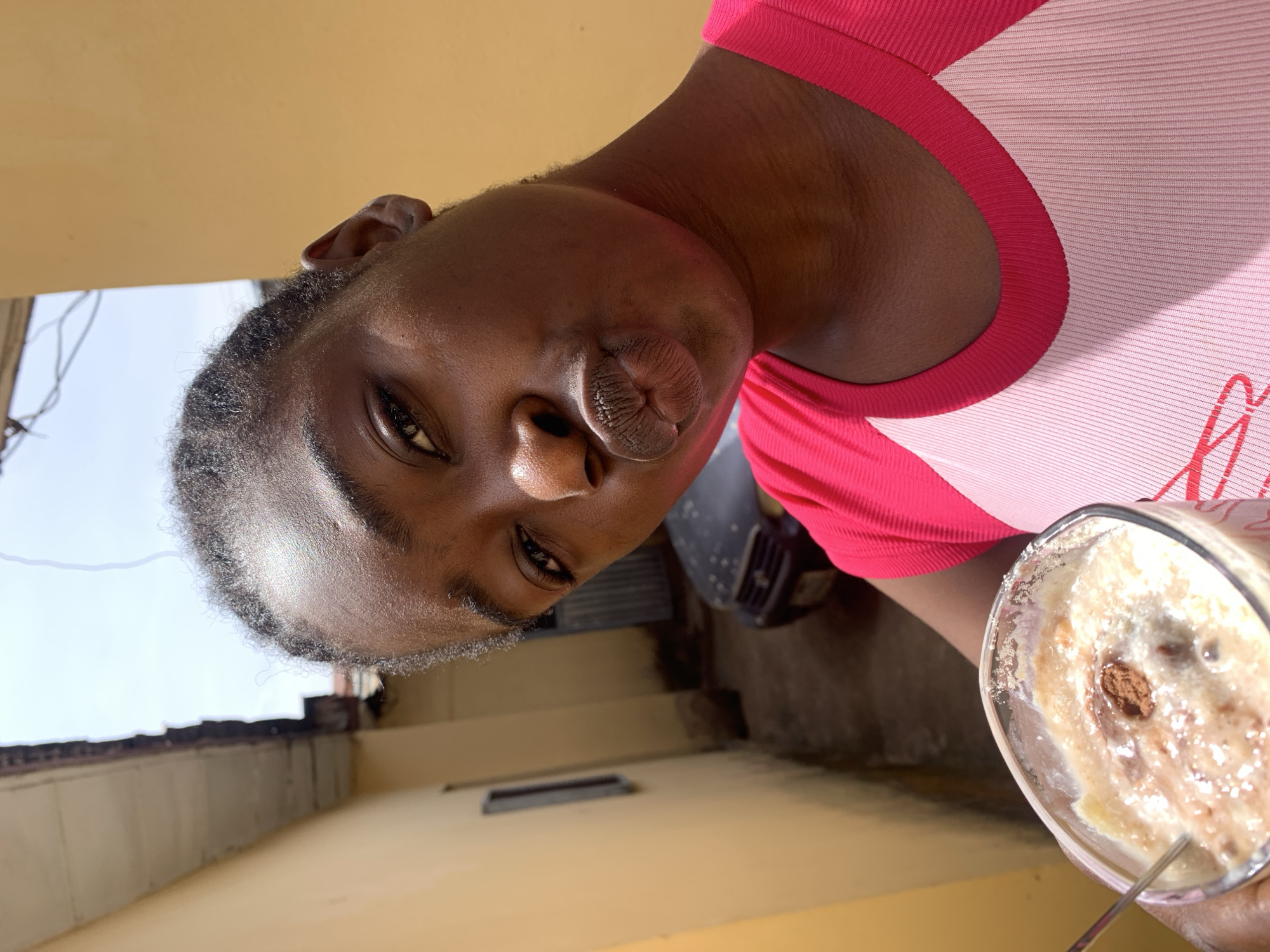 A person is indoors in Rafin Par, Nigeria, holding a glass of a blended, iced beverage. The person is wearing a pink and white ribbed top and has dark skin. Their hair is dark and short. The background includes a beige wall and a glimpse of outdoor elements, possibly rooftops, under a bright, clear sky. There is also a glimpse of a vehicle behind the person.