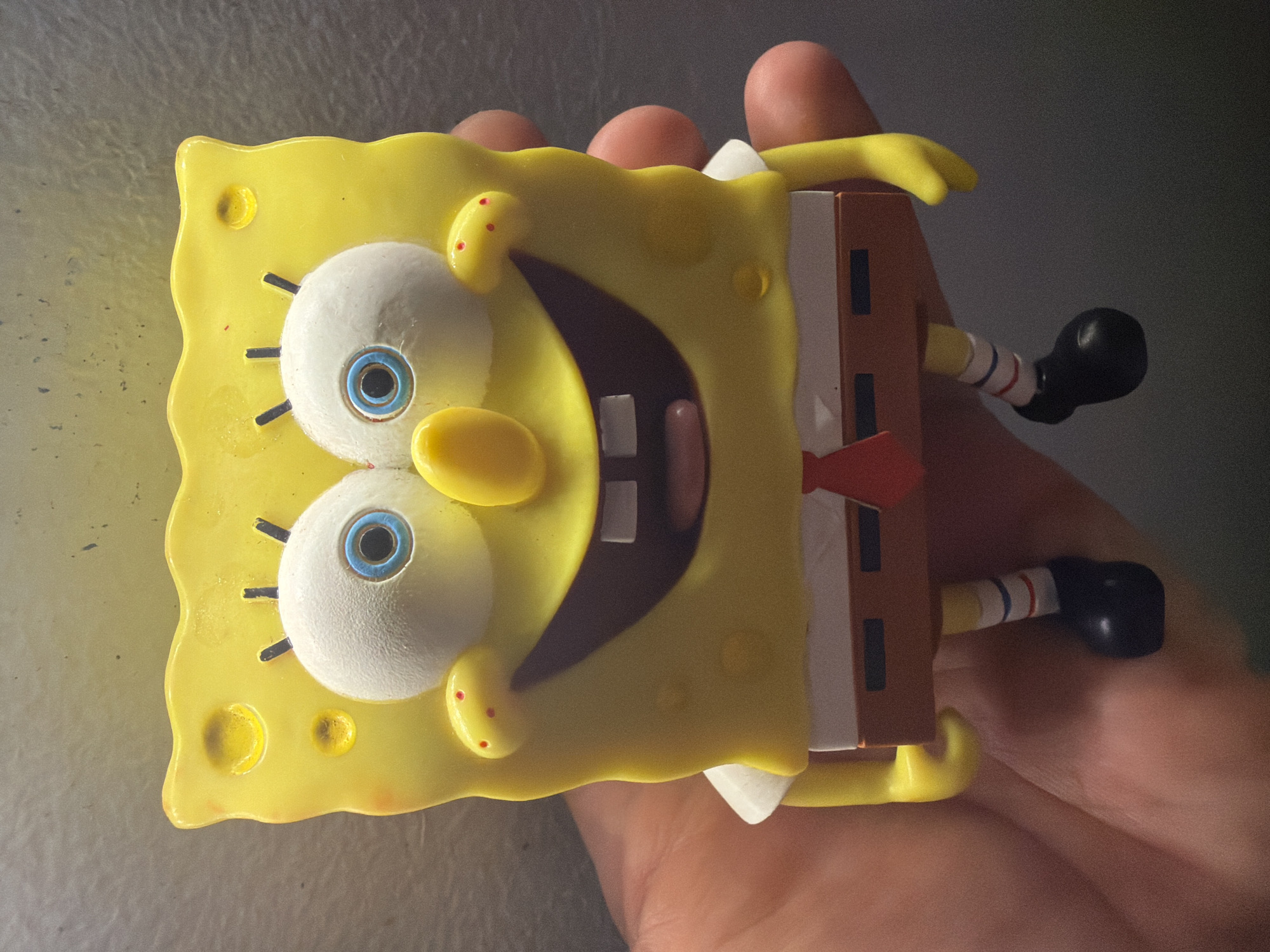 A close-up, low-angle shot shows a Spongebob Squarepants toy figure held in a hand. The toy is bright yellow and features Spongebob's signature square shape, large white eyes with blue irises, a yellow nose, and a wide, smiling mouth with two white teeth visible. He is wearing his characteristic white shirt, brown pants, and a red tie. His legs are visible, with striped socks and black shoes. The background is a dark, mottled surface, possibly a refrigerator or wall. The hand holding the toy is slightly out of focus. There are no identifiable city or environmental landmarks.
