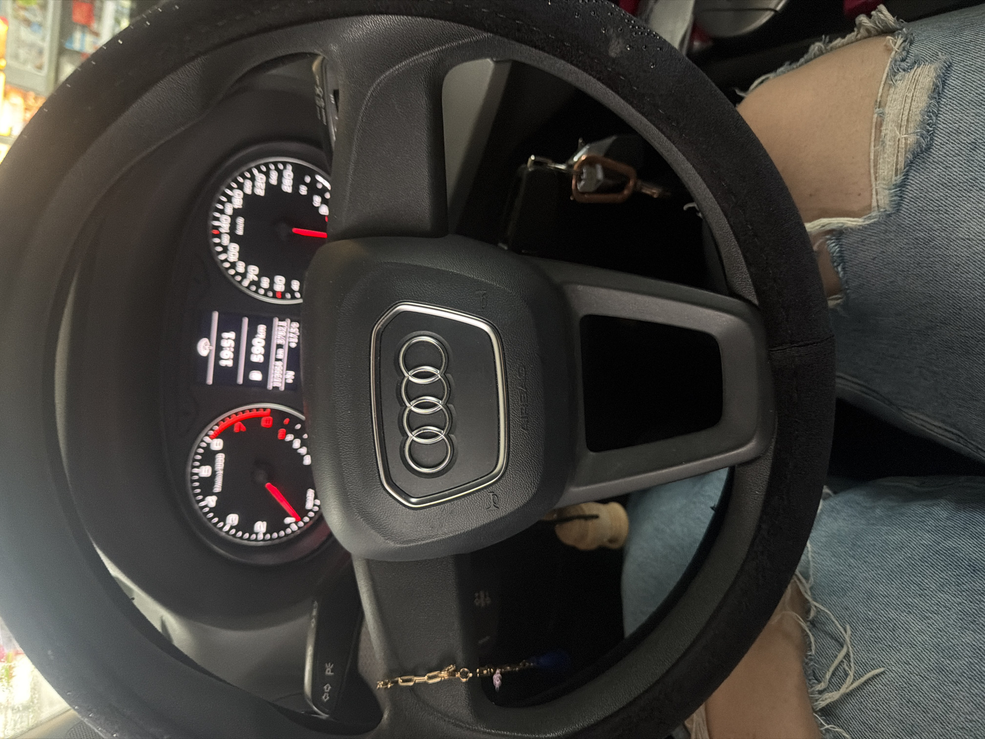 The image shows the interior of a car, specifically the steering wheel of an Audi. The instrument panel is visible behind the steering wheel, displaying information such as speed, RPM, and possibly other vehicle data. Keys are in the ignition. Part of a person's leg is visible on the right side of the image, clad in ripped denim jeans. A gold bracelet is dangling in the car. The location is unknown.