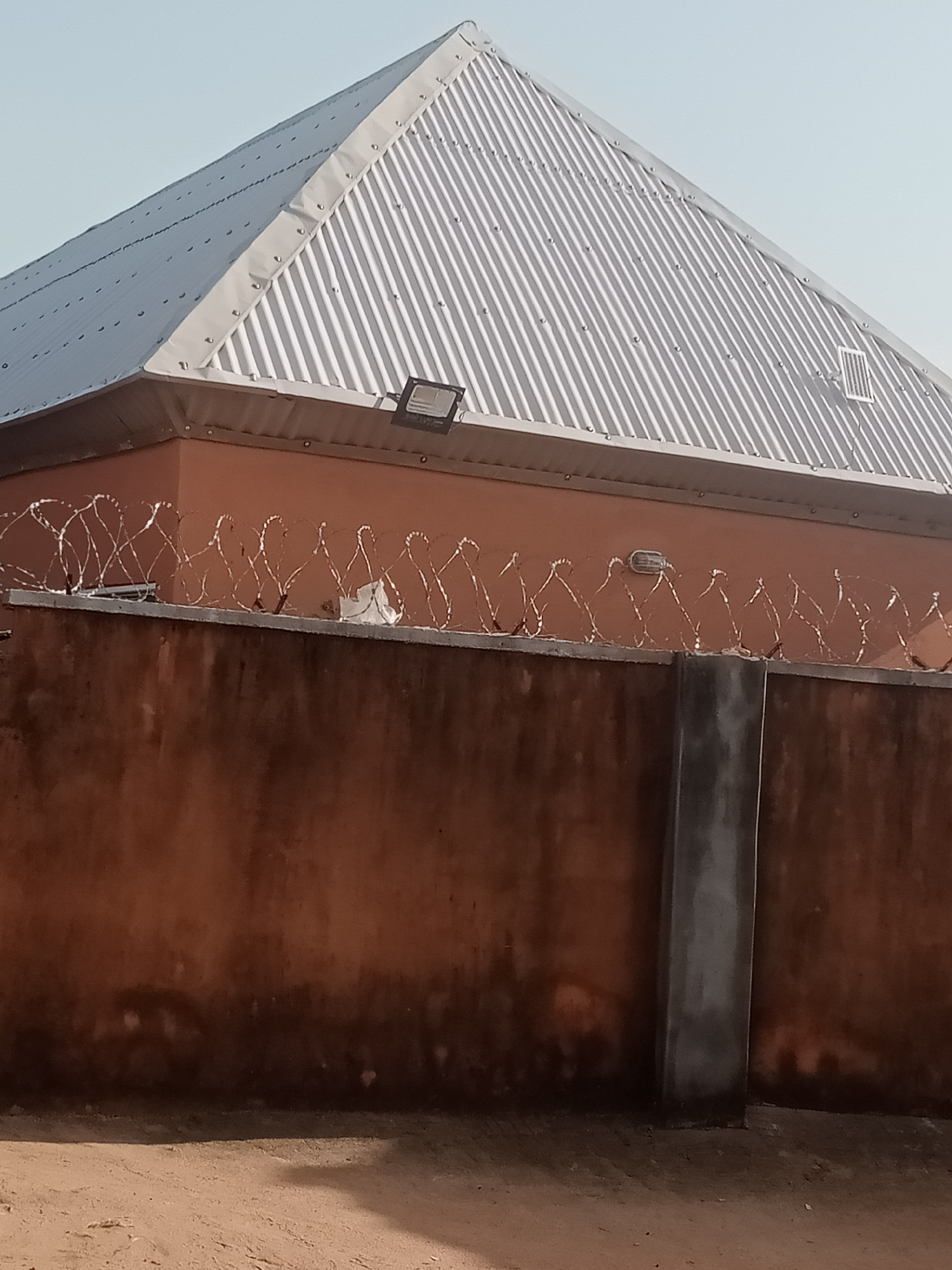 The image shows a section of a building in Jalingo, Nigeria, characterized by a corrugated metal roof and an orange-colored wall. Razor wire is visible along the top of the wall, indicating a security measure. A concrete pillar supports the wall. The ground in front of the wall is covered with sand and dirt. The sky is clear and light blue, suggesting it is daytime. There are no people or animals visible in the image. The overall scene depicts an exterior view of a property in what appears to be a semi-urban or rural setting.