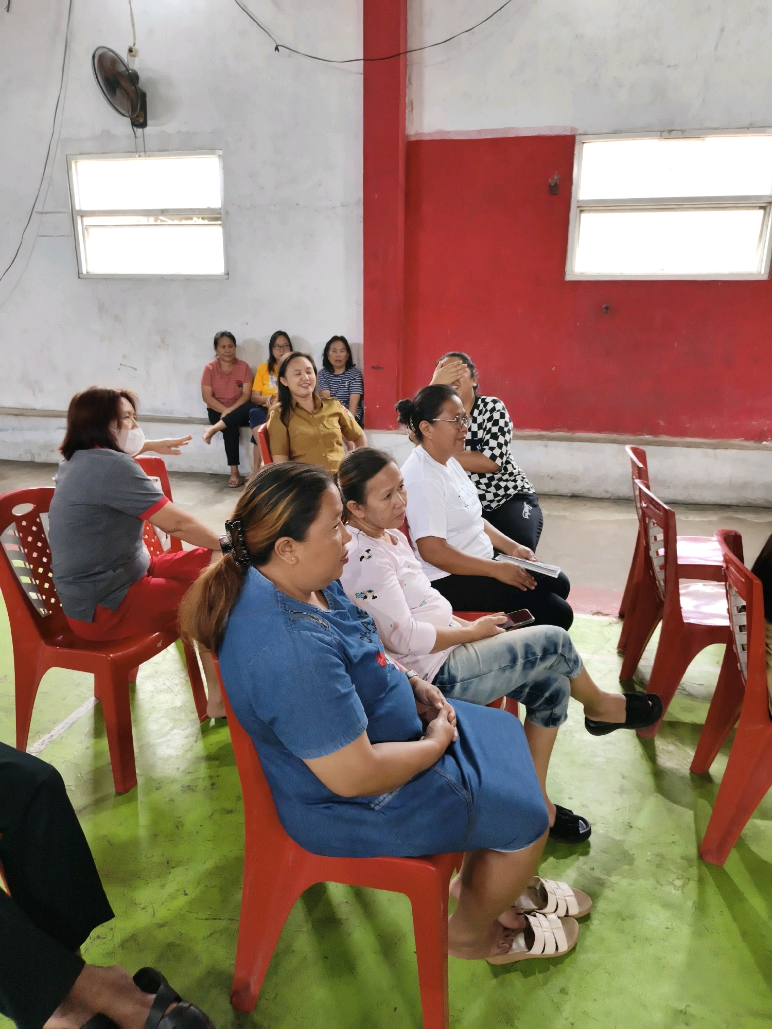 In Tombatu Satu, Indonesia, a group of women are seated on red plastic chairs in what appears to be a community hall or large room. The walls are painted white and red, with two windows letting in bright, natural light. A ceiling fan is mounted on the white wall. The floor is a vibrant green.
In the foreground, a woman in a blue denim dress is seated on a red chair, looking to her right. She has her hair tied back with a black clip. To her left, another woman in a light pink shirt and jeans is seated, holding a mobile phone in her hand and looking forward. Next to her, a woman wearing glasses and a white t-shirt is also seated, looking towards the front and holding a folded document. Behind her, a woman with a checkered shirt is partially visible, covering her face with her hand.
Further back, a woman in a brown shirt is seated, smiling and looking to her right. To her left, a woman in a yellow shirt is seated, looking forward. Next to her, a woman in a striped shirt is seated. In the far back left, a woman in a grey t-shirt and red pants, wearing a white face mask, is seated and gesturing with her left hand. The red plastic chairs are arranged in rows, and some are empty. The overall impression is a gathering or meeting in progress.