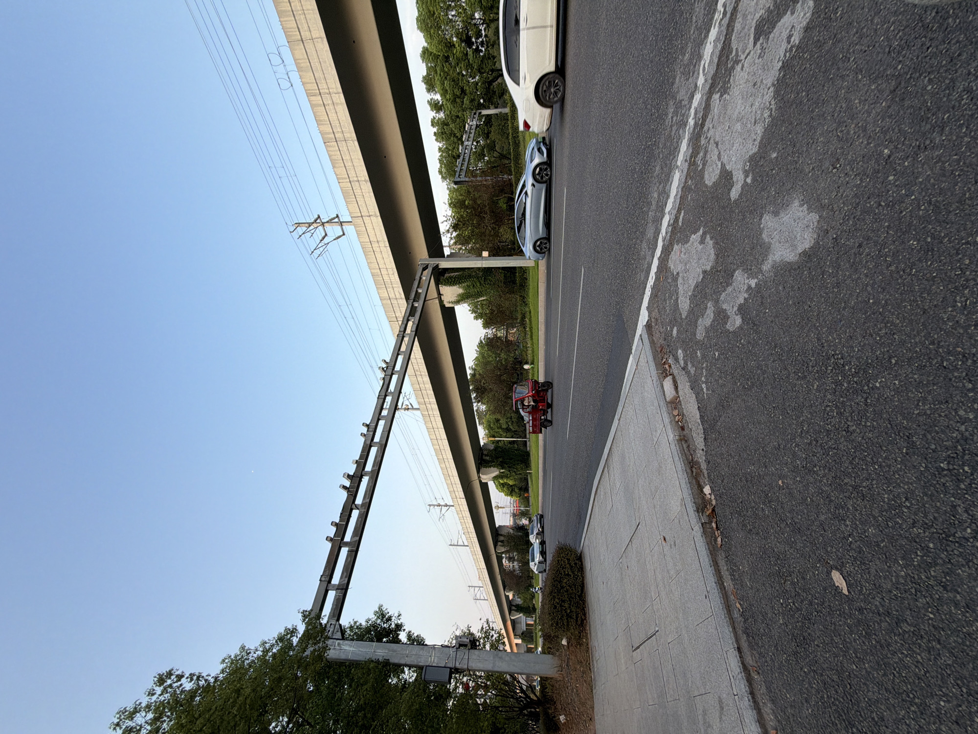 This image depicts a street scene in Jiangjin District, Chongqing, China, near an elevated roadway or overpass. The sky is clear and blue, suggesting daytime. On the street, there are several vehicles: a white car on the right, a silver car in the middle, and a red three-wheeled vehicle with a canopy, driven by a person, moving towards the left. A sidewalk runs along the left side of the road, made of grey paving stones, with a green bush bordering it. Overhead, a large concrete structure, likely part of an elevated transport system, spans across the scene. Power lines are visible on this structure and beyond. Trees and foliage line the sides of the road, with some greenery climbing the supports of the overpass. The overall environment is urban, with infrastructure for transportation and natural elements.