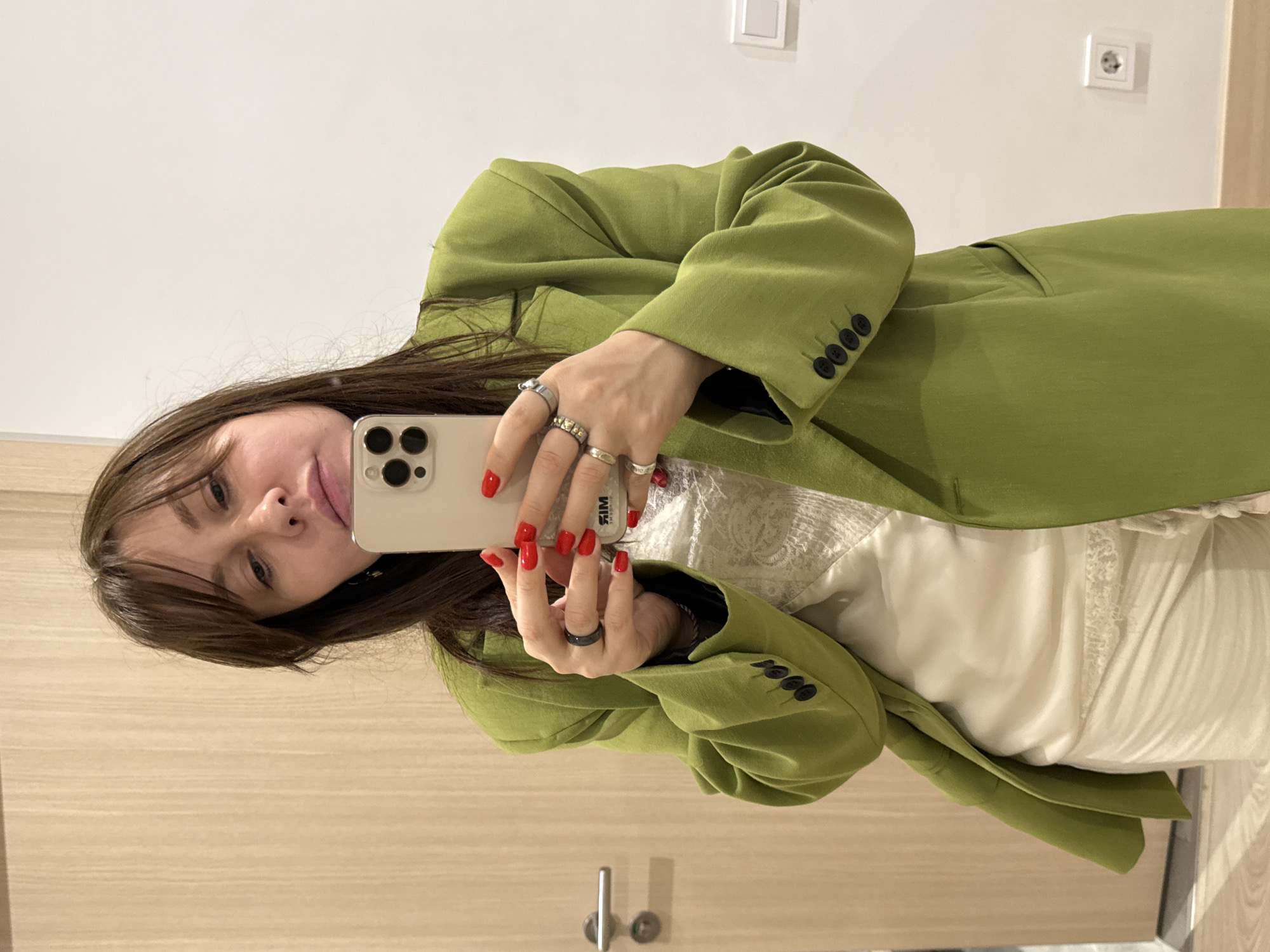 A woman is taking a selfie indoors. She is wearing a light green blazer over a white dress with lace details. She has brown hair with bangs. She is holding a smartphone in her hands, which have red nail polish and multiple rings. The background shows a light-colored wooden door, a white electrical outlet, and a white switch on the wall. The location is likely not discernible as Moscow, Russia from the visual data available.