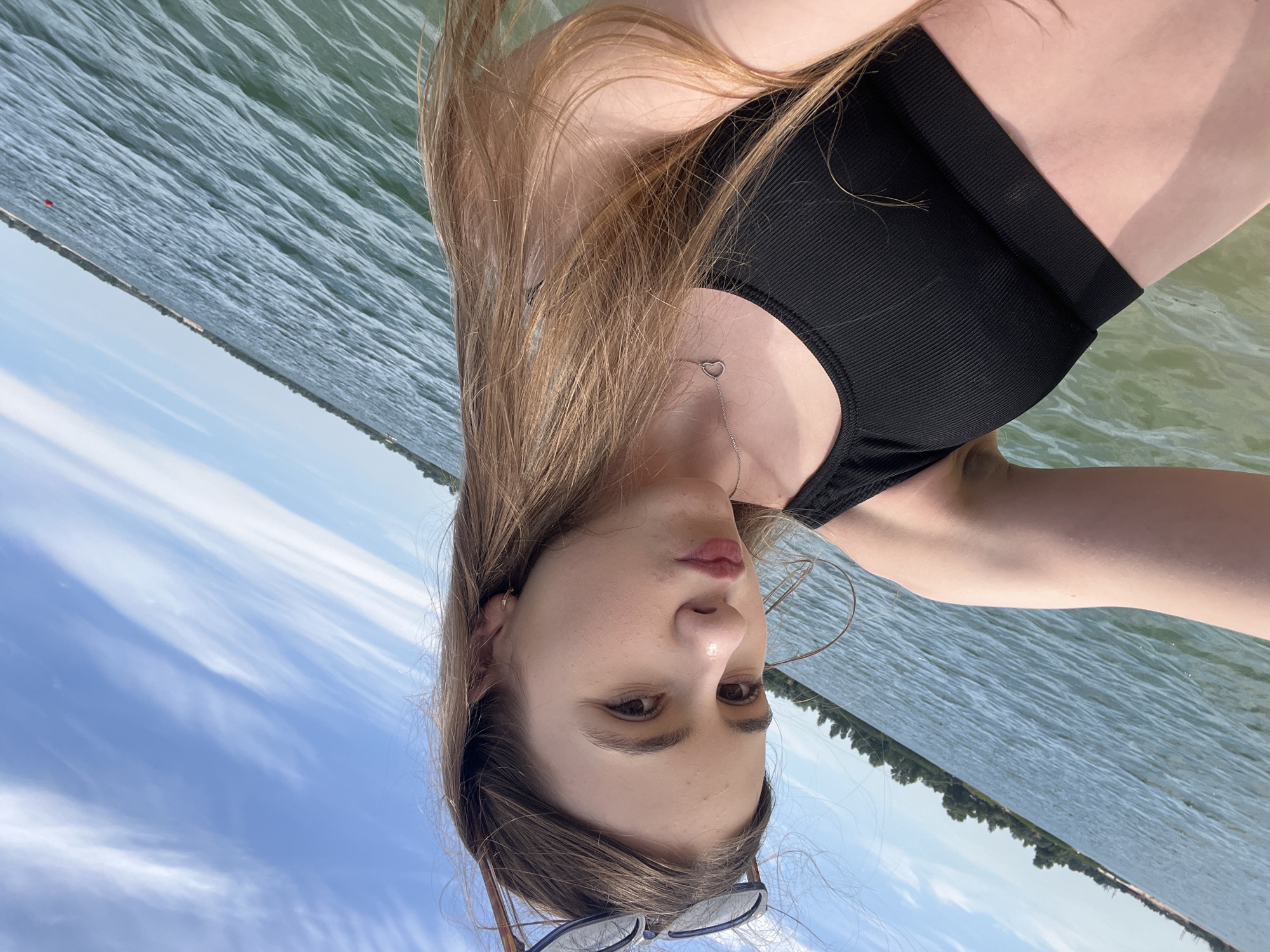 The media file shows a close-up, low-angle, over-the-shoulder shot of a young woman in a black bikini top, submerged in water. The image is oriented sideways. Her long, light brown hair is spread out, partially covering her face. She is looking towards the camera with a neutral expression. A delicate silver necklace with a heart pendant is visible on her chest. The background consists of rippling water and a blue sky with wispy white clouds. In the distance, a tree line is visible along the horizon. The lighting suggests it is daytime, likely sunny, with the sun reflecting off the water. The presence of water and the bikini top indicate the activity is likely swimming or enjoying a day at the beach or lake. The location is identified as Yelyzavetivka, Ukraine. There is no visible text in the media file.