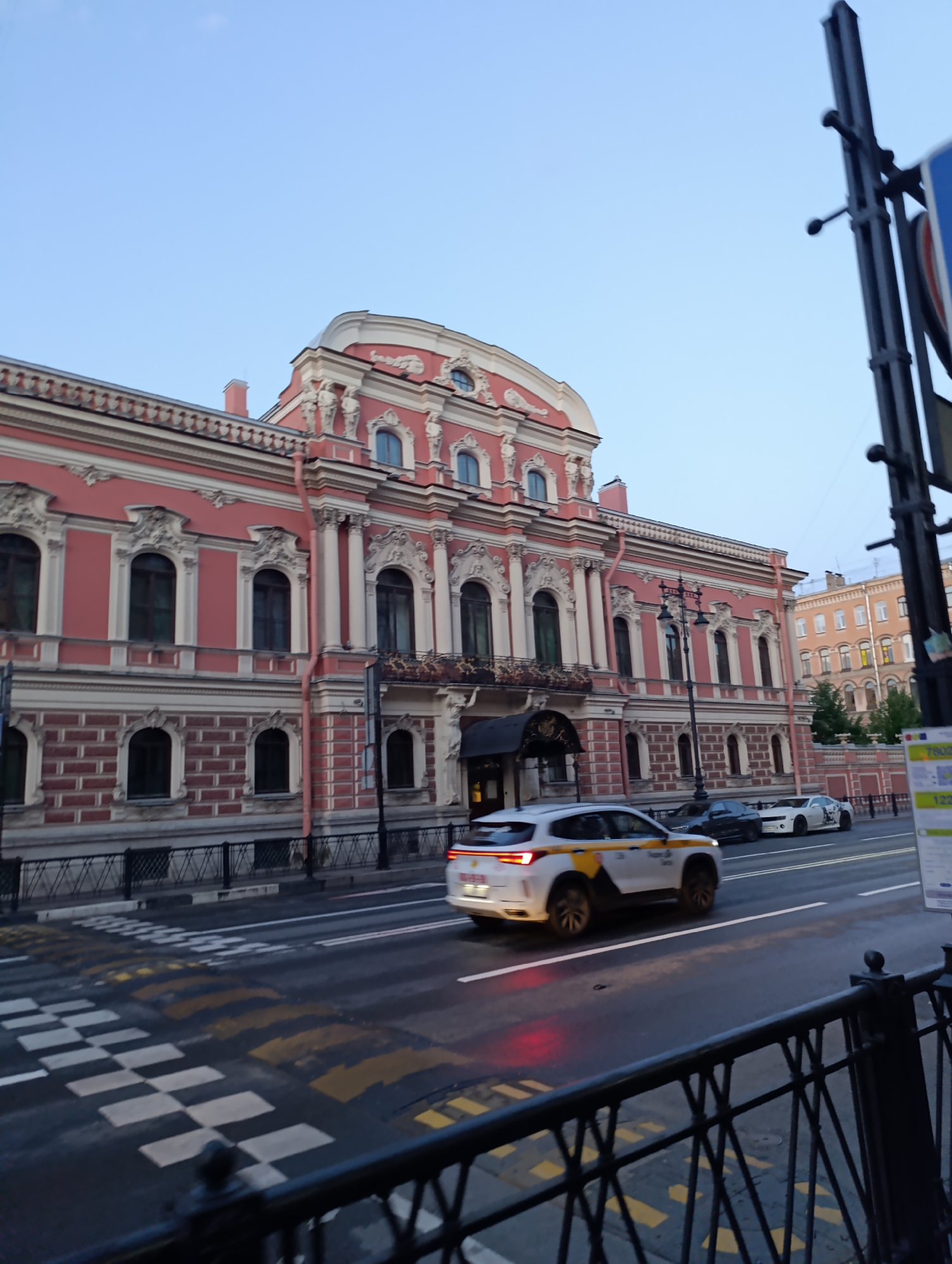 A white SUV with yellow and orange markings, likely a taxi, drives across a wet street with a checkered pedestrian crossing. Behind it, two cars, a black sedan and a white sports car, are also on the road. The scene is set in front of a grand, pink building with intricate architectural details, including arched windows and decorative columns. The building's facade is adorned with white molding and brickwork patterns.
Further in the background, other buildings with lighter facades are visible, suggesting an urban environment. A lamppost stands near the sidewalk. The sky is a clear, pale blue, indicating it is likely early morning or late evening. The wetness of the road suggests recent rain. The overall impression is a city street in Russia, possibly Saint Petersburg, during a quiet time of day. No people are clearly discernible.
Visible text includes "122" on a sign on the right side of the image and partial text on the taxi.