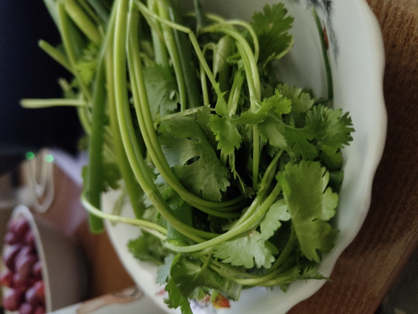 A close-up, eye-level, indoor shot shows a pile of fresh green cilantro and green onions on a white, scalloped-edge plate. The plate has a subtle floral pattern with hints of red and black. The cilantro leaves are delicate and lobed, while the green onions are thicker and have long, slender stems. The items are arranged loosely, with stems and leaves overlapping. In the background, out of focus, is a white bowl filled with red beans, and a dark object with two faint green lights. The scene is set on a wooden table.