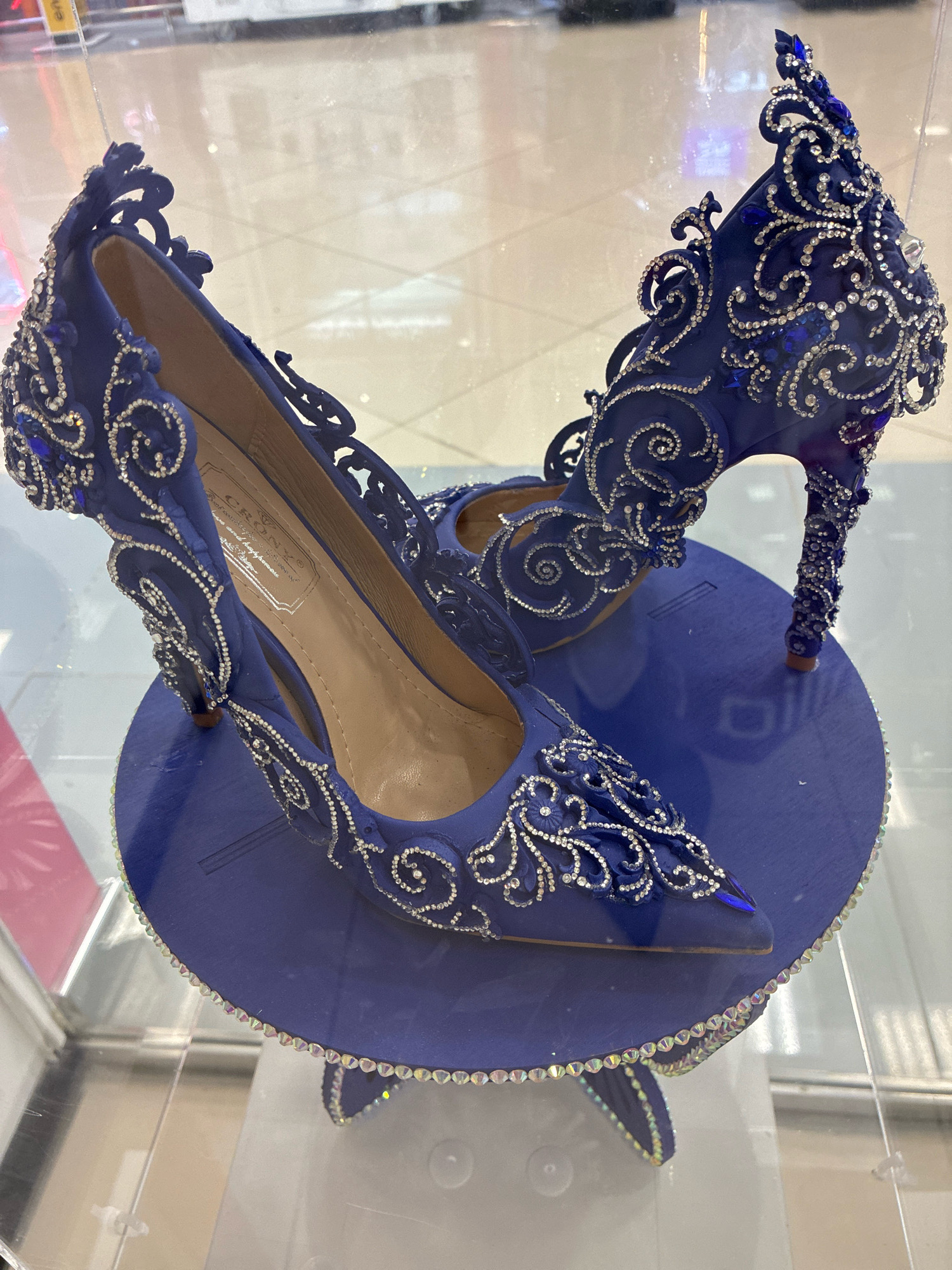 A pair of ornate, royal blue high-heeled shoes are displayed on a matching blue circular stand. The shoes are heavily embellished with silver and clear rhinestones arranged in swirling patterns and floral motifs. Some larger, faceted blue stones are also incorporated into the design. The interior of the shoes is a light tan color and a label is visible inside the left shoe. The stand itself is also decorated with a border of iridescent rhinestones. The shoes and stand appear to be displayed within a glass case, possibly in a retail environment, with a blurred background that suggests a shopping mall or similar public space. The image was captured in Moskovskiy, Russia.