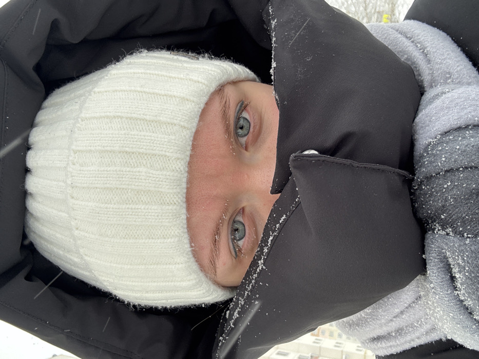 The image presents a close-up, frontal view of an individual outdoors in Nizhnekamsk, Russia, during snowy conditions. The subject's face is largely covered by winter attire, revealing light-colored skin, blue eyes, and the upper forehead. They are wearing a white ribbed knit beanie, and a black winter jacket with the hood drawn around their head. A section of the jacket's collar or an integrated neck covering is fastened across the lower face with a silver-colored snap, protecting the chin and mouth area. A grey and black patterned scarf is visible below this black fabric, wrapped around the neck. Small flakes of snow are present on the subject's eyebrows, the black jacket material, and the scarf. The background indicates an urban environment with a light-colored building partially visible to the left and snow on the ground. Individual snowflakes are perceptible in the air, confirming active snowfall.