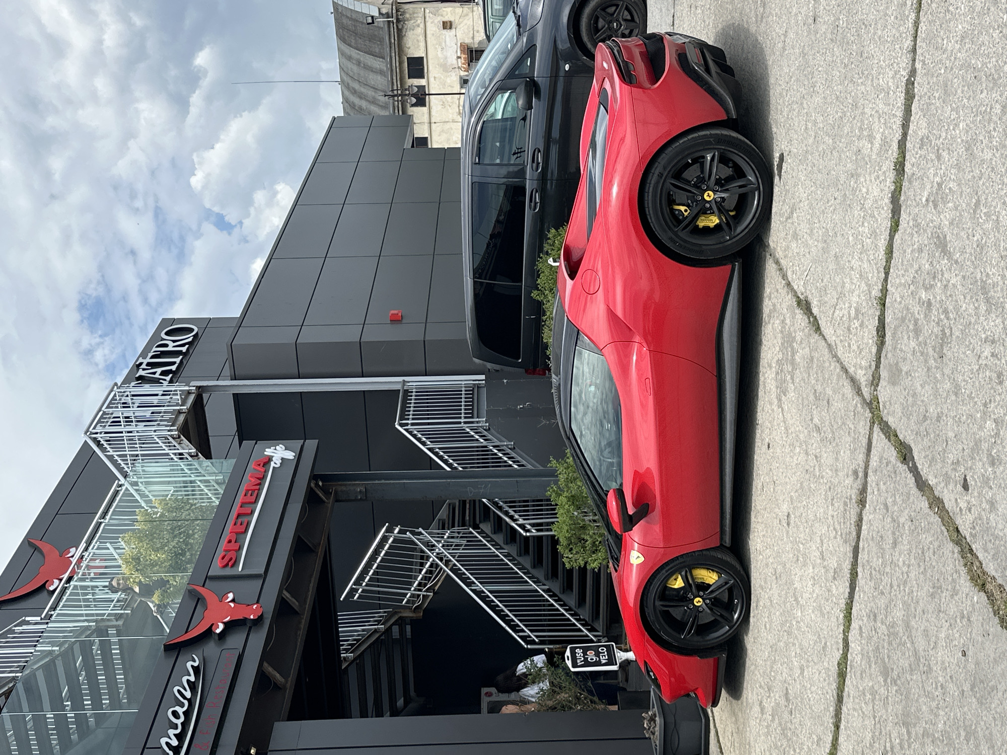 The image shows a red Ferrari parked on a concrete surface in front of a modern building in Varna, Bulgaria. The building has a black facade with metal staircases leading to the upper floors. A sign for "Spetema" and "mamm & fish Restaurant" is visible on the building's exterior. A black van is parked behind the Ferrari. The sky is cloudy. The scene appears to be outdoors. A small sign is visible near the base of the building.