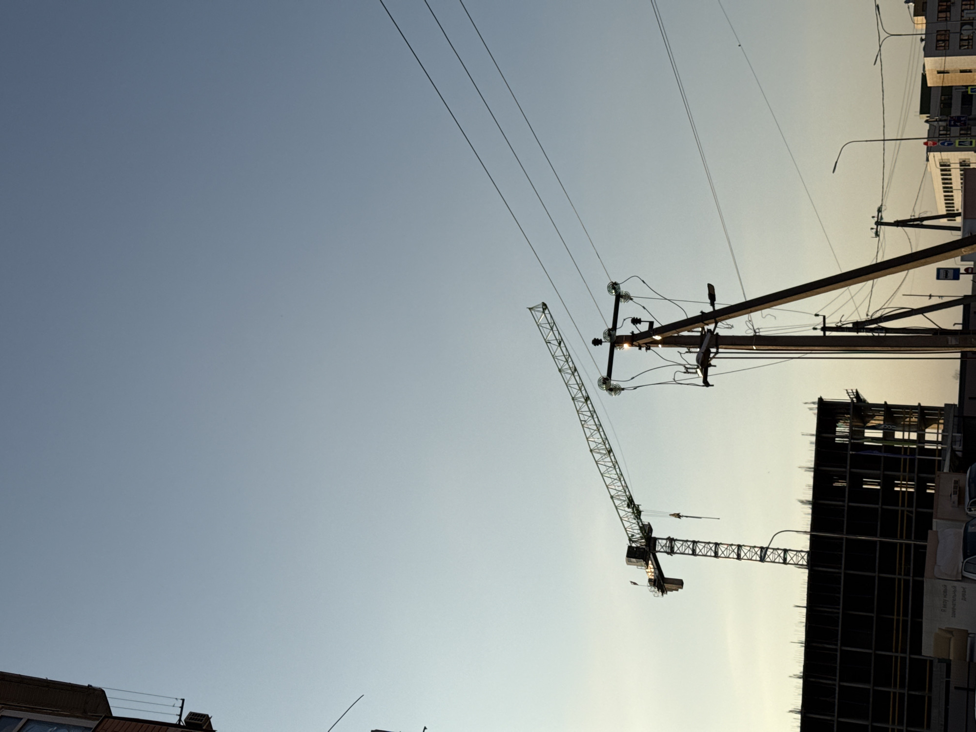 The image depicts a construction site in the city of Unknown, Unknown, under a clear sky with the horizon showing a slight gradient in color. A tall construction crane dominates the midground, positioned next to a building under construction, which appears as a skeletal frame. Electrical poles and wires run across the upper portion of the frame. Other buildings are visible in the background along with street lighting.