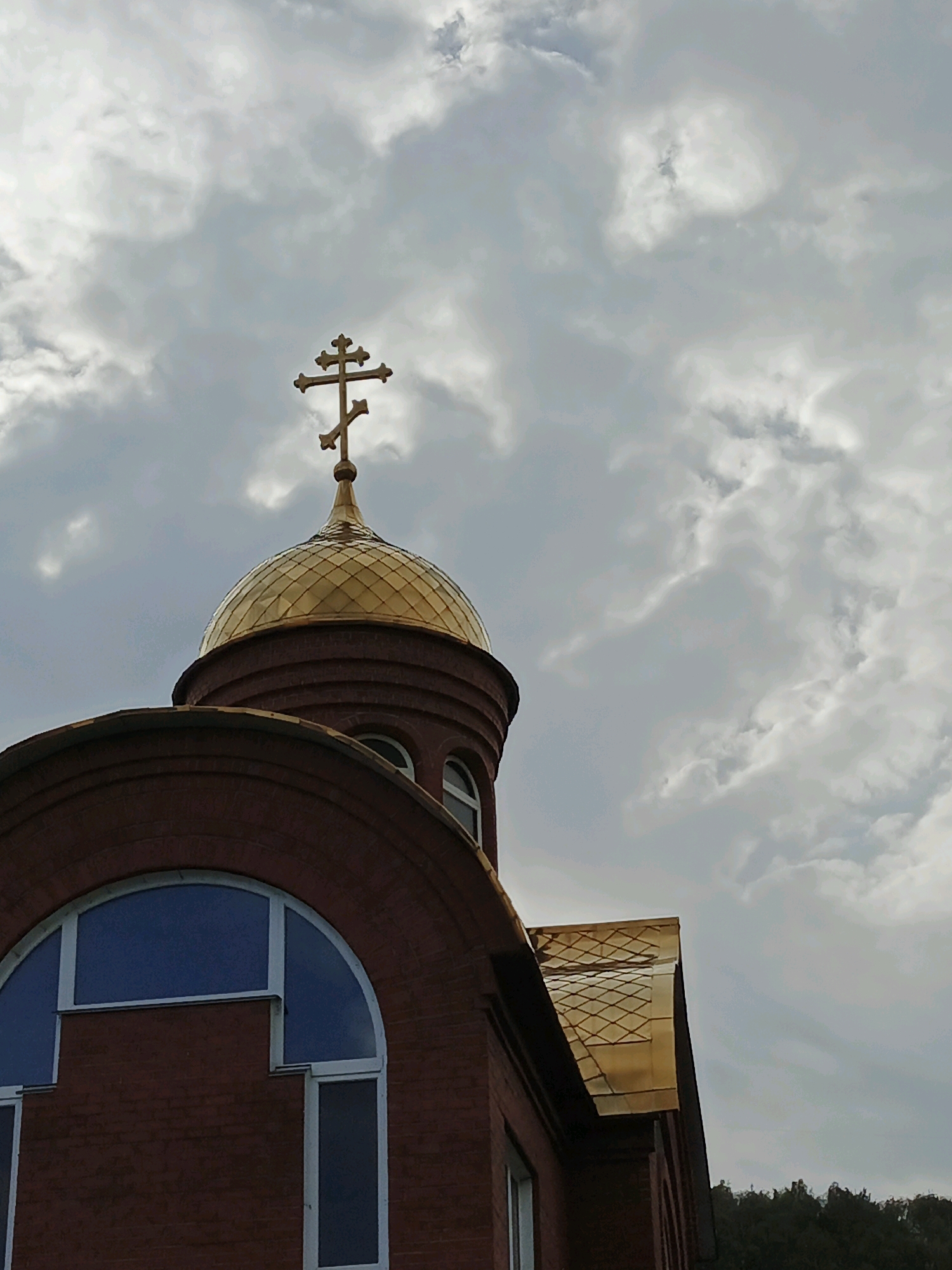 This is a low-angle, medium shot of a brick building with a golden dome and cross against a cloudy sky. The building appears to be a church, specifically the Church of the Holy Trinity in Zlatoust, Russia. The brickwork is red and textured, with arched windows. The dome is a prominent golden color and is topped with a golden cross. The sky is overcast with dramatic clouds. In the background, the dark green tops of trees are visible. No people are present in the image.