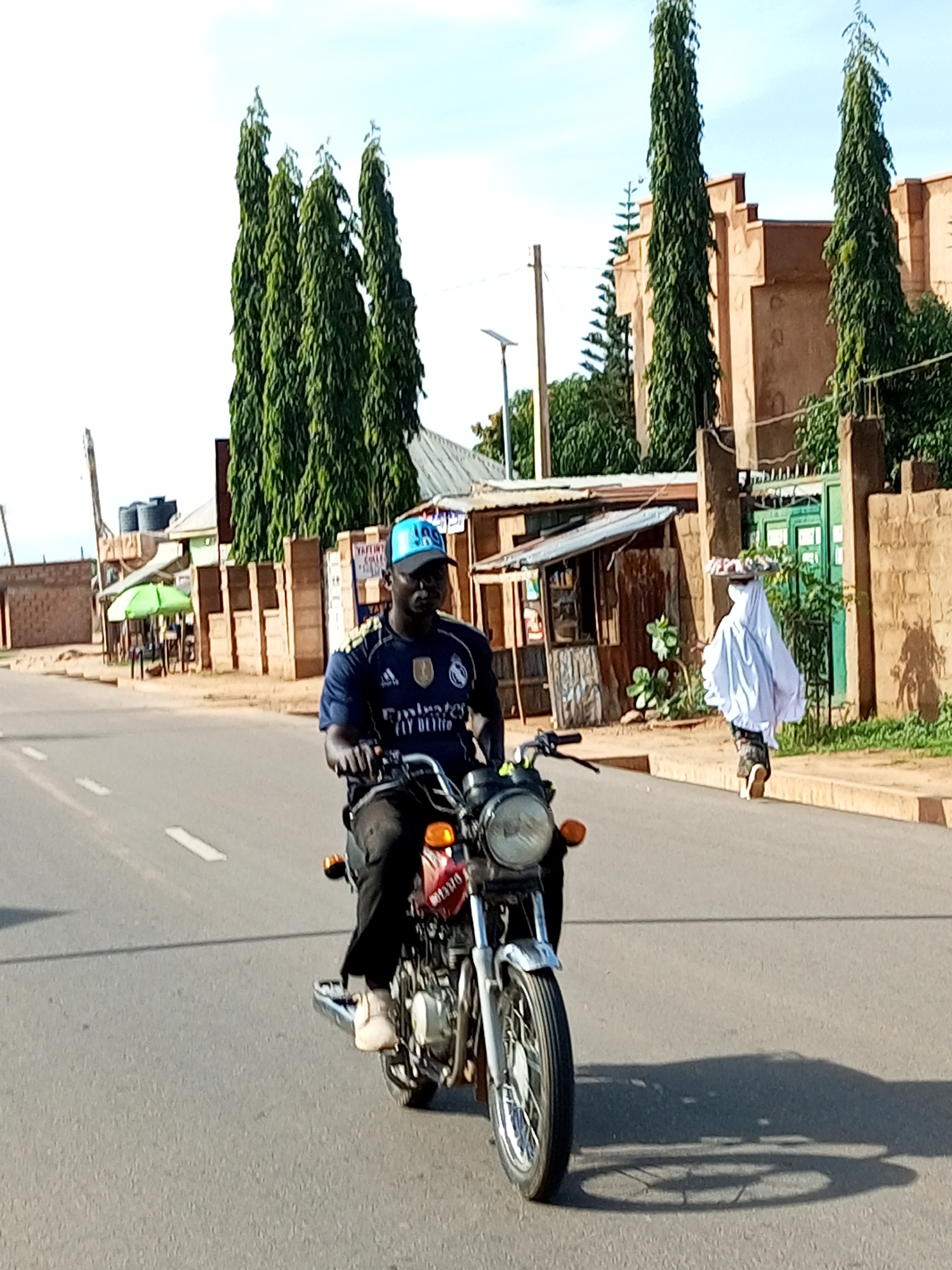 This is a street-level, medium-shot photograph taken in Tempure, Nigeria. A man wearing a blue Real Madrid jersey and a blue baseball cap is riding a red and silver motorcycle down a paved road. He is looking forward, and his hands are on the handlebars. In the background, a person draped in a white cloth is walking away from the camera, carrying a tray on their head. The scene is lined with buildings made of earth-toned brick and some green trees. The sky is partly cloudy.