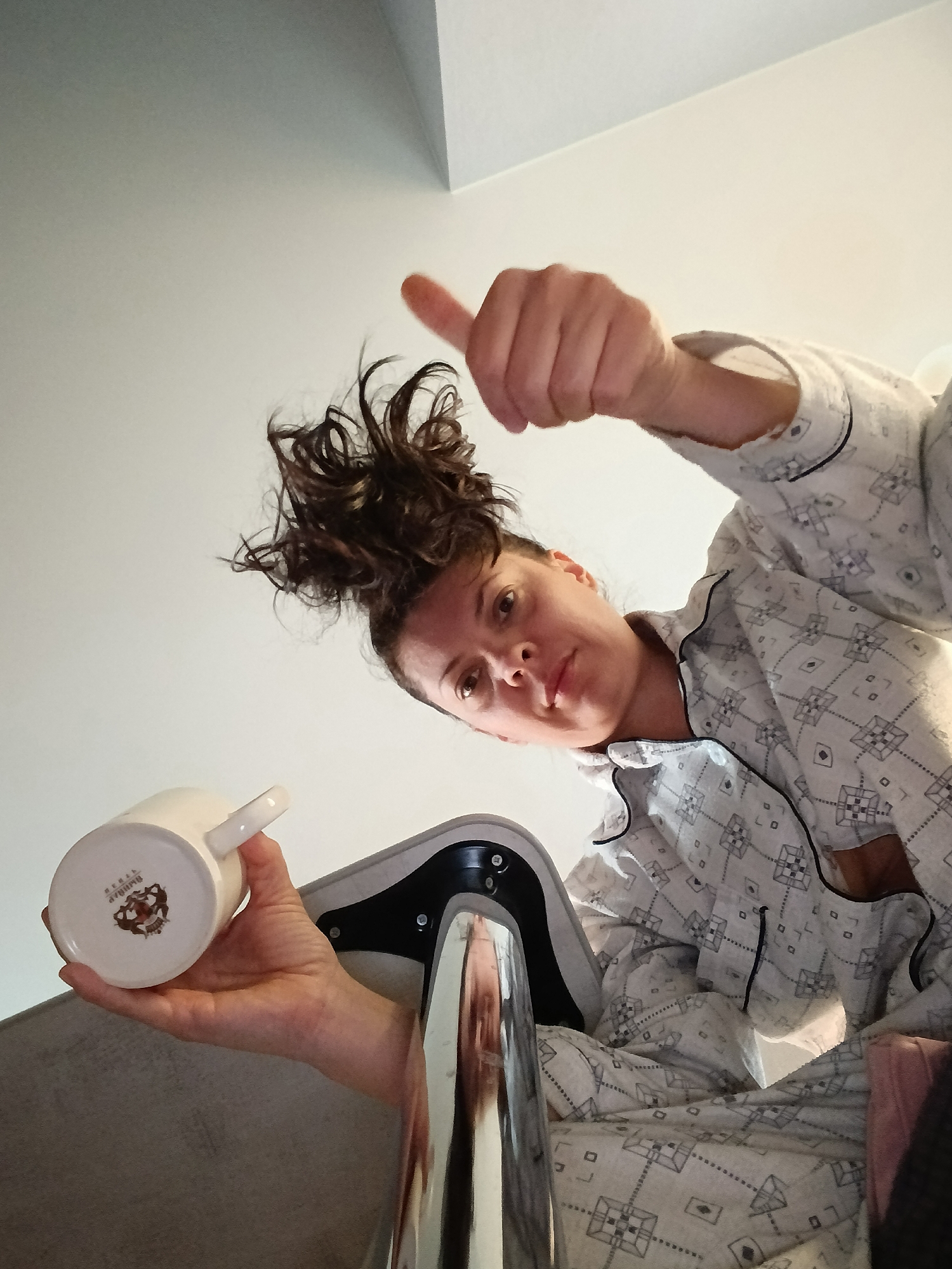 In Saratov, Russia, a woman wearing a patterned pajama set is captured in a low-angle shot, holding a white mug and giving a thumbs-up gesture. Her dark, curly hair is styled in a messy bun on top of her head. The background consists of a plain white wall, suggesting an indoor setting, possibly a bedroom. The woman's facial expression is focused, and she appears to be looking directly at the camera. The mug she holds has a small emblem on its base, and her right hand is extended forward with her thumb raised in a positive sign. The overall scene is casual and intimate, with a focus on the woman and her gesture.