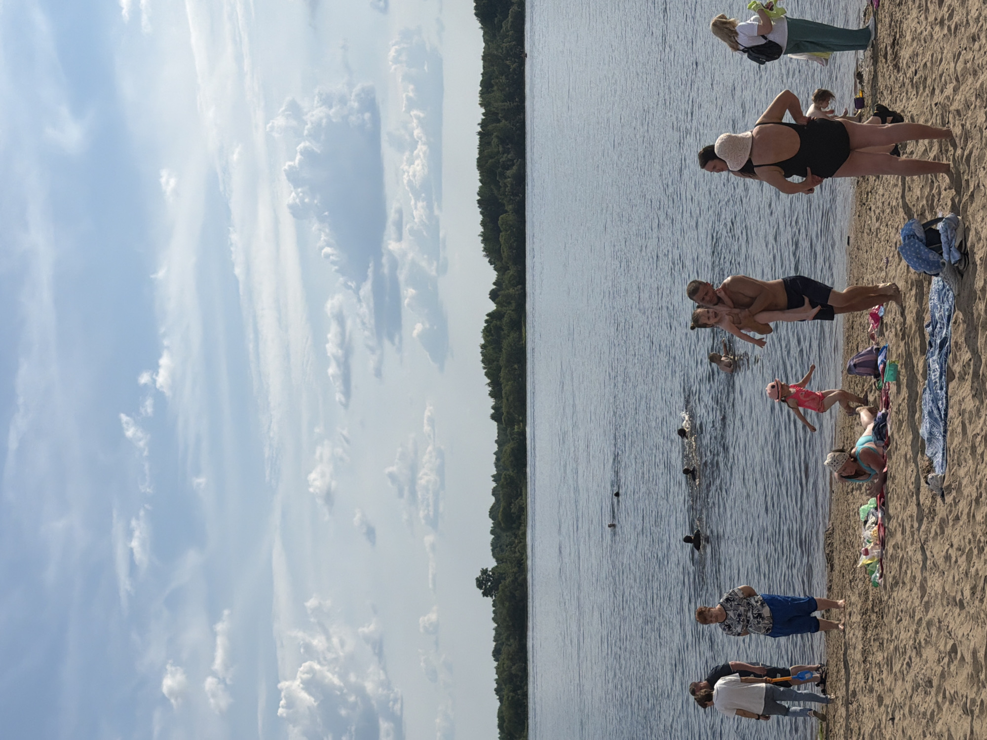 In Solikamsk, Russia, on a sunny day, a group of people are enjoying themselves at a beach by a wide river. The sandy shore is dotted with several individuals and families. Some people are in the water, swimming and playing, while others are relaxing on towels laid out on the sand. A man is holding a young child, and a woman in a black swimsuit is adjusting her attire. Children are playing on the beach, and various beach accessories like bags and towels are scattered around. In the background, the river stretches towards a densely wooded shoreline under a partly cloudy sky.