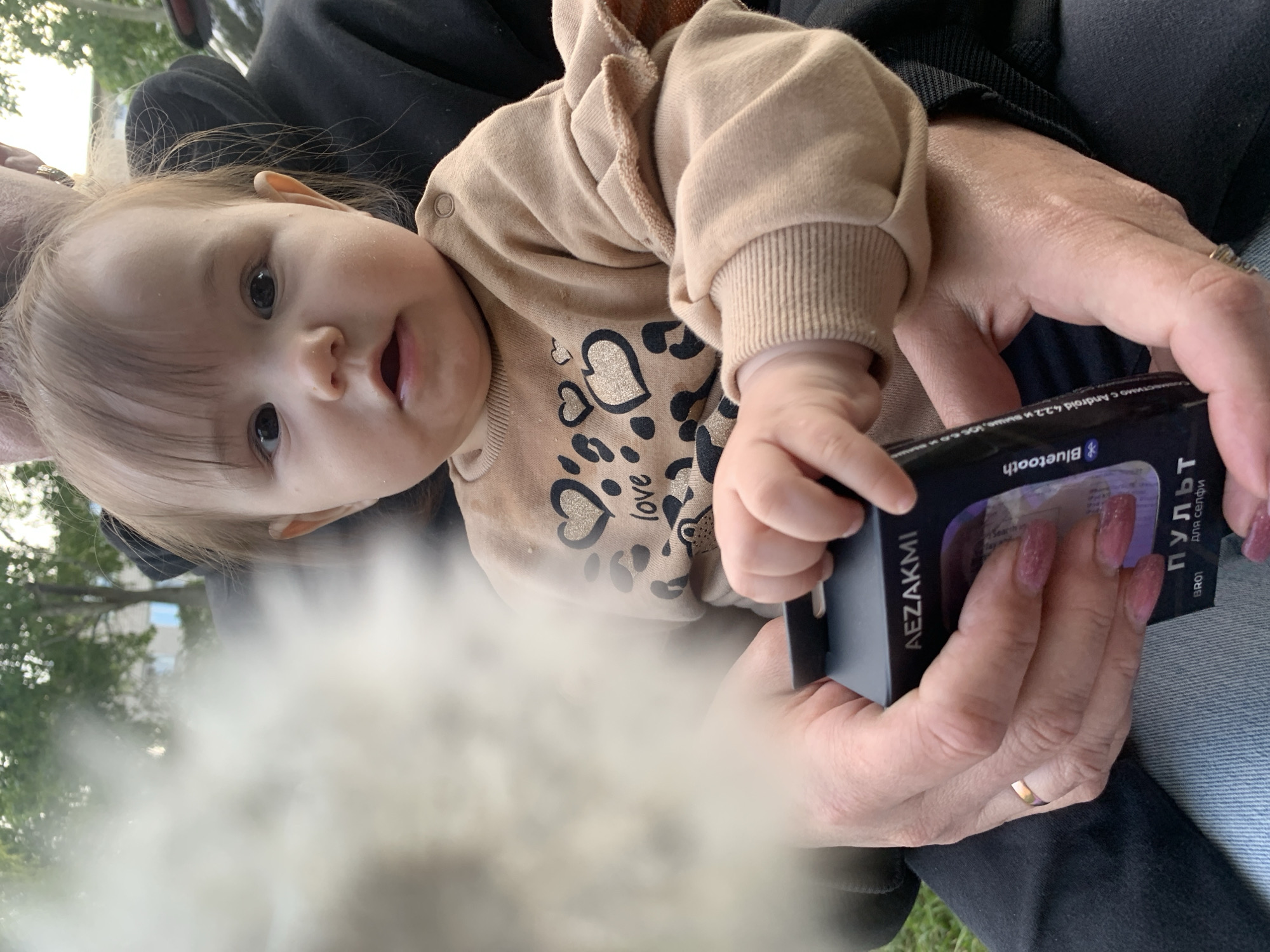 A baby wearing a tan-colored hooded sweatshirt with heart and leopard print lies in someone's arms, partially holding a black box labeled "AEZAKMI Bluetooth пульт для селфи BR01," which translates to "remote for selfie." The adult's hands hold the box, revealing manicured nails and a gold ring. The location is unknown, but the text on the box is in Russian, and the name of the manufacturer, AEZAKMI, is associated with Chaikovsky, Russia. The background is slightly blurred, showing greenery and suggesting an outdoor setting.