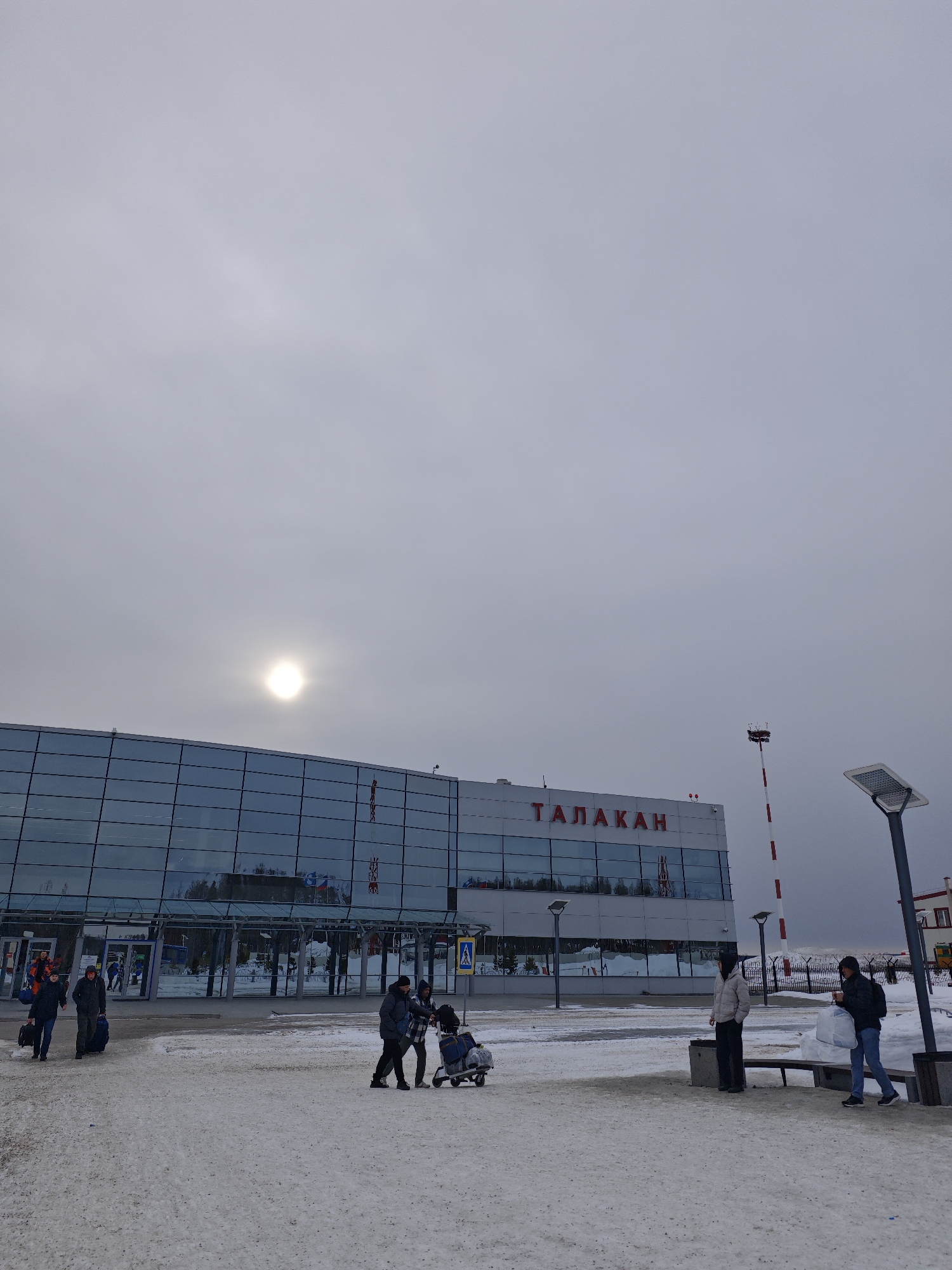 The image depicts the exterior of the Талакан airport in Талакан, Russia on a cloudy day. The building is a two-story structure with a glass facade and the name of the city in red letters above the entrance. There is snow on the ground. Several people are present, some pulling luggage carts, others standing near benches. A striped red and white communication tower stands to the right of the building. Streetlights are visible in the foreground and background.