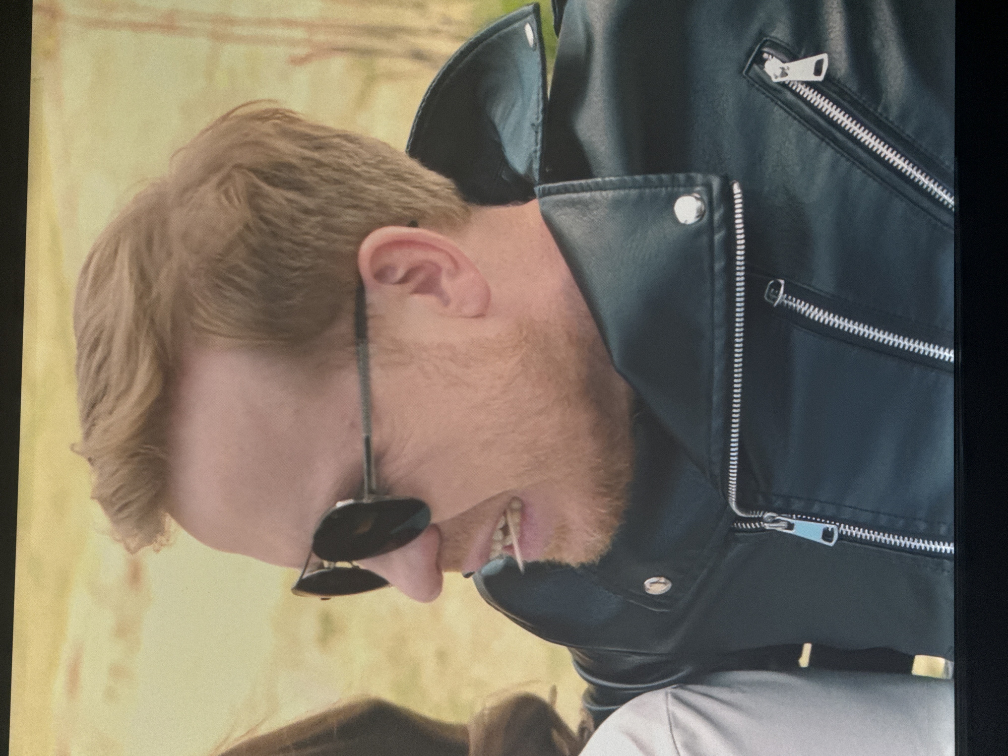 The image shows a close-up, profile view of a man with reddish-blonde hair, wearing aviator sunglasses, a black leather jacket with zippers, and what appears to be a toothpick or similar object in his mouth. The man is looking to his left, with his mouth open in an expression that could be interpreted as speaking or grimacing. The background is a blurred, yellowish-orange, suggesting an outdoor setting during daylight. There is a partial view of another person, likely a woman, to the left of the frame, whose shoulder and hair are visible. The focus is on the man's face and upper body. There is no information in the image that would indicate the location in the city of Saratov, Russia.
