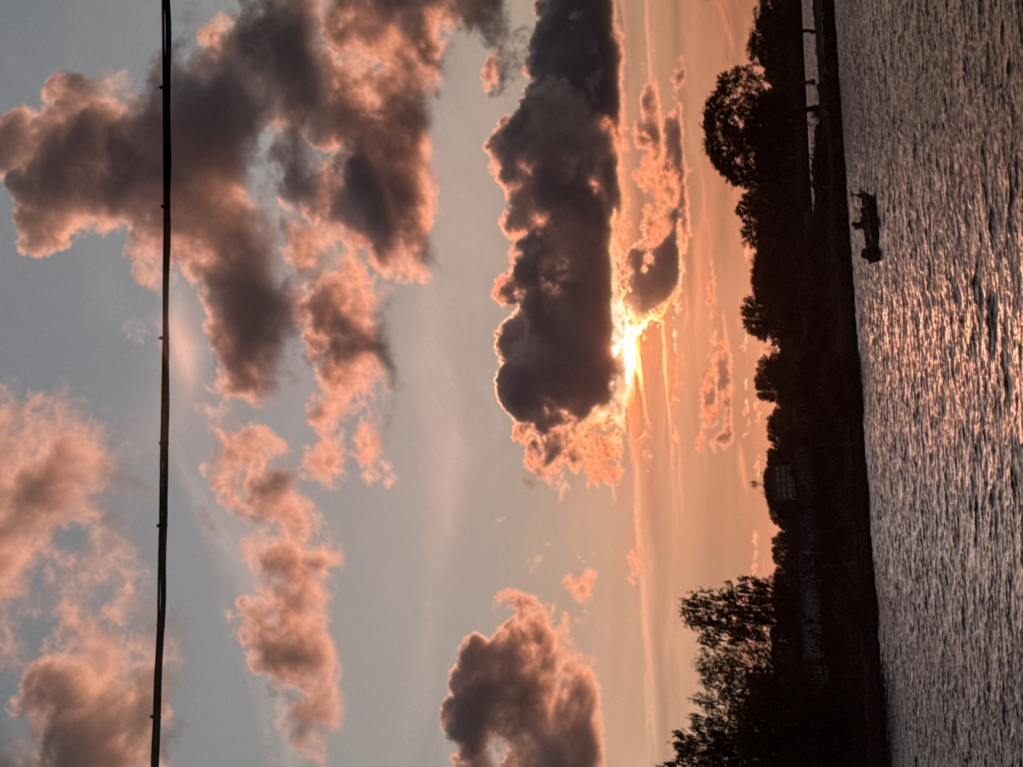 The image depicts a sunset over a body of water, likely the Daugava River, in Riga, Latvia. The sky is filled with dramatic clouds, tinged with hues of pink and orange from the setting sun. A silhouetted treeline marks the distant shore, with some buildings and structures faintly visible within the silhouette. In the foreground, the water's surface reflects the warm colors of the sky. A small motorboat with at least one person is visible in the water, moving slowly. A power line stretches horizontally across the upper portion of the image.
