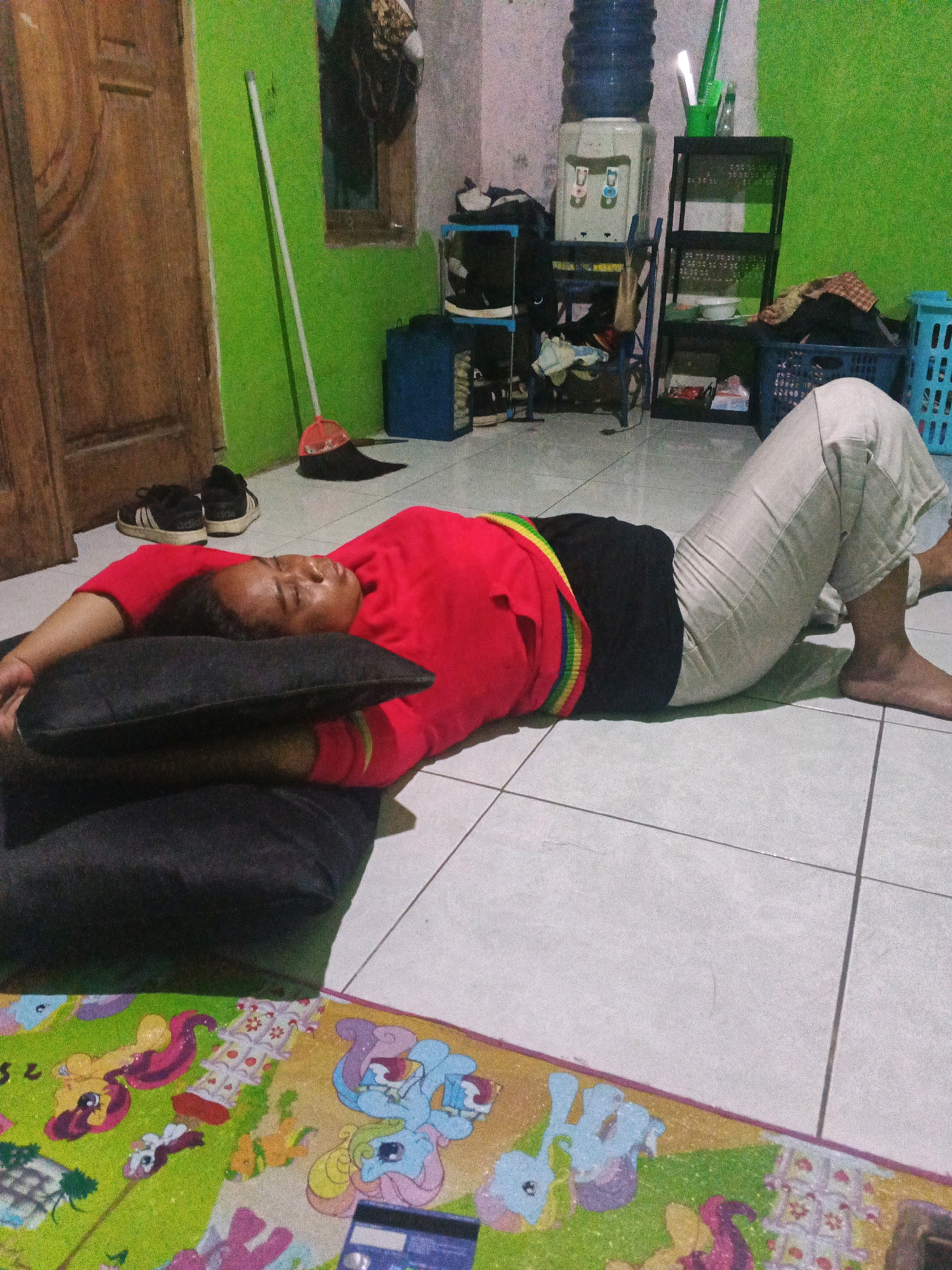 A person is lying on their back on a tiled floor in what appears to be a room in Semarang, Indonesia. They are resting their head on a dark pillow, with their arms outstretched. The individual is wearing a red shirt and light-colored pants. In the background, there are various household items, including a water dispenser, shelving units with items, a broom, a wooden door, and laundry baskets. A colorful mat with cartoon characters is visible on the floor in the foreground.