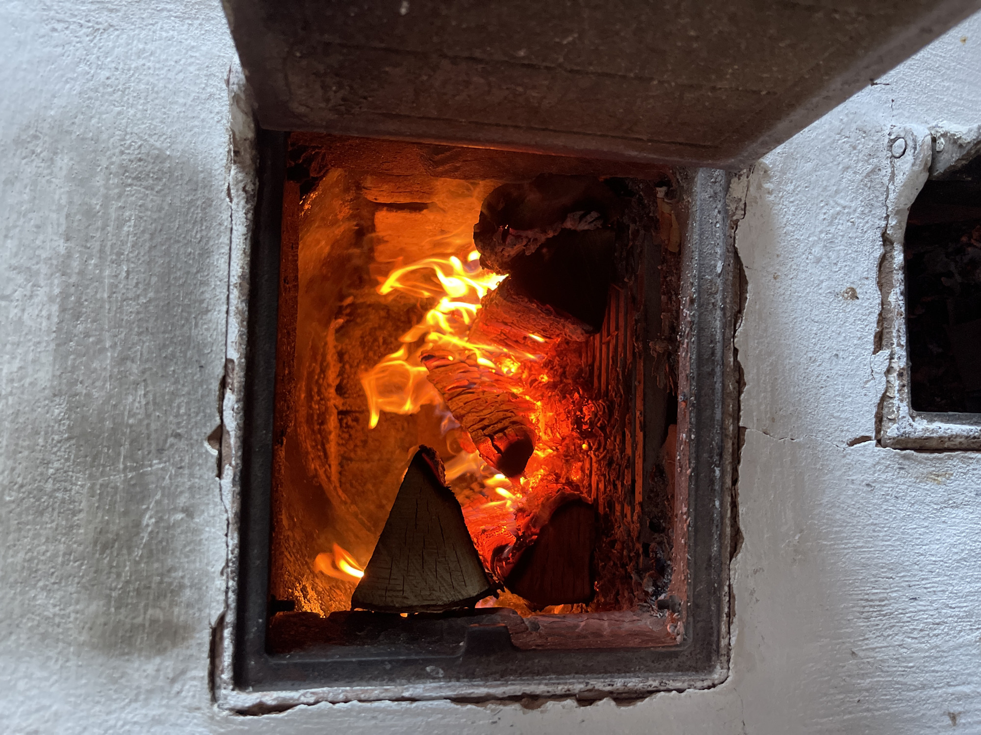 The image shows the interior of a wood-burning oven. The oven is constructed of painted white masonry, with a dark metal frame around the opening. Flames are visible, consuming pieces of wood arranged inside. A portion of the oven door is open, revealing the burning wood. To the right of the open oven door, another small opening is visible in the masonry. The exact location within the city of Zhigalki, Russia cannot be determined from the image.