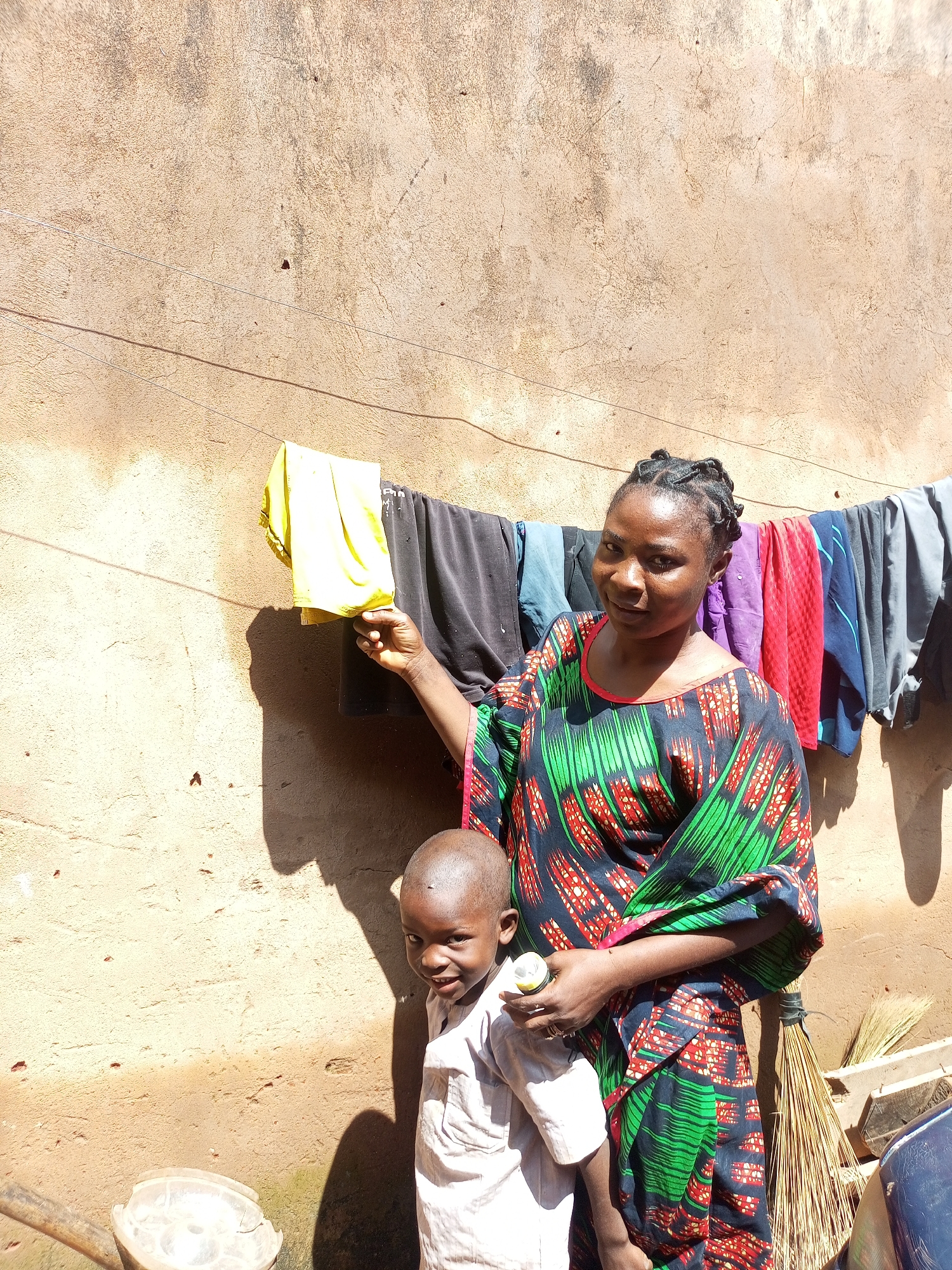 In a sun-drenched outdoor scene in Tempure, Nigeria, a woman and a young child stand against a textured, earthen wall. The woman, wearing a vibrantly patterned dress with green, red, and black motifs, has her hair styled in intricate braids and holds a small, white cylindrical object in her right hand. Her gaze is directed towards the camera. Beside her, a young boy in a light-colored shirt smiles at the camera, his left hand resting on a transparent plastic container. Clothes, a mix of colors and textures including a bright yellow garment, are hanging on a clothesline strung across the wall behind them. A broom made of natural fibers leans against the wall to the right of the woman. The ground in the foreground appears to be dirt.