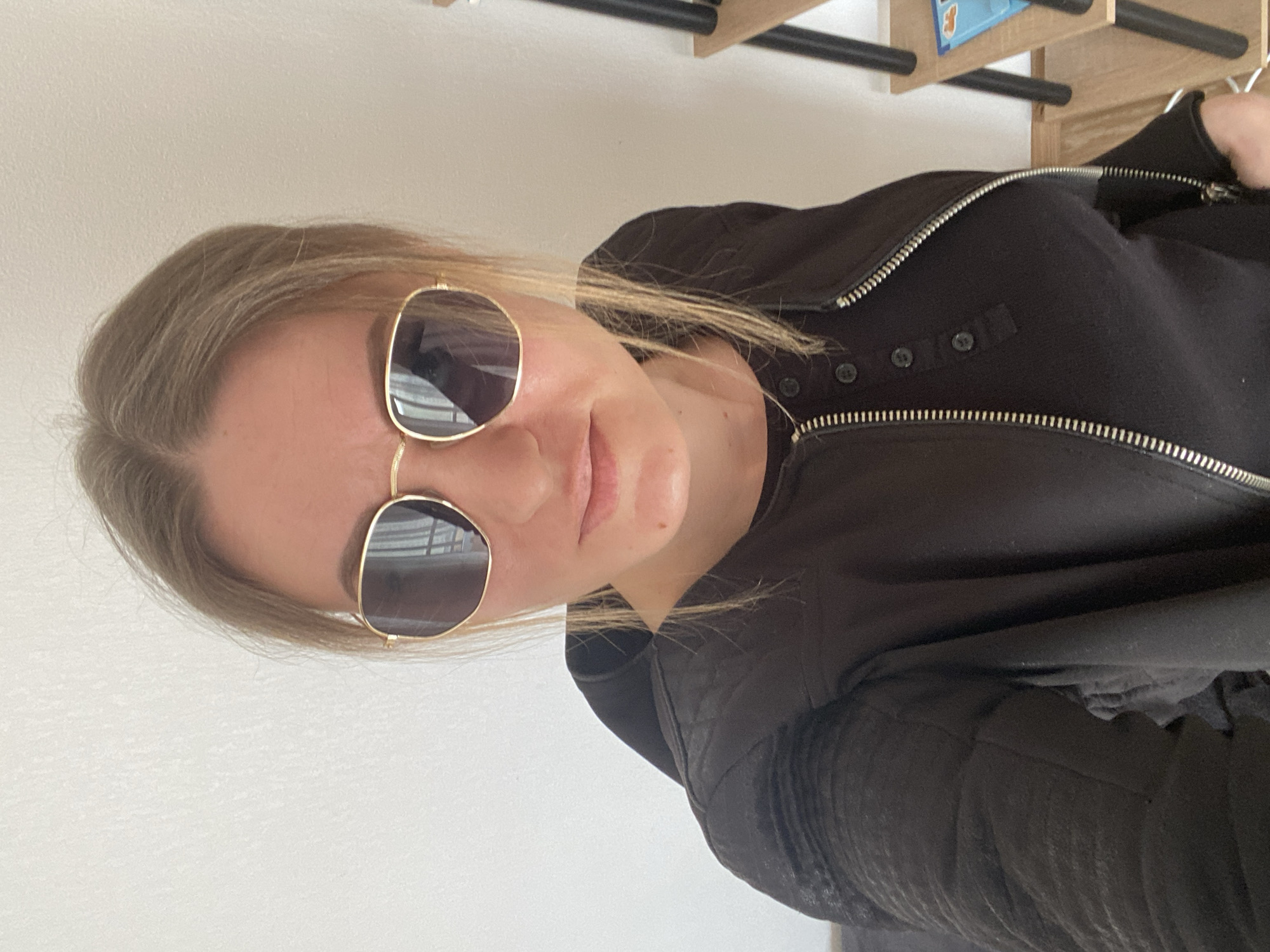 A woman with blonde hair and sunglasses is wearing a black zippered jacket. She appears to be indoors, against a white wall. In the upper portion of the image, a wooden structure with black metal supports is visible. The scene gives no indication of being located in Saint Petersburg, Russia.