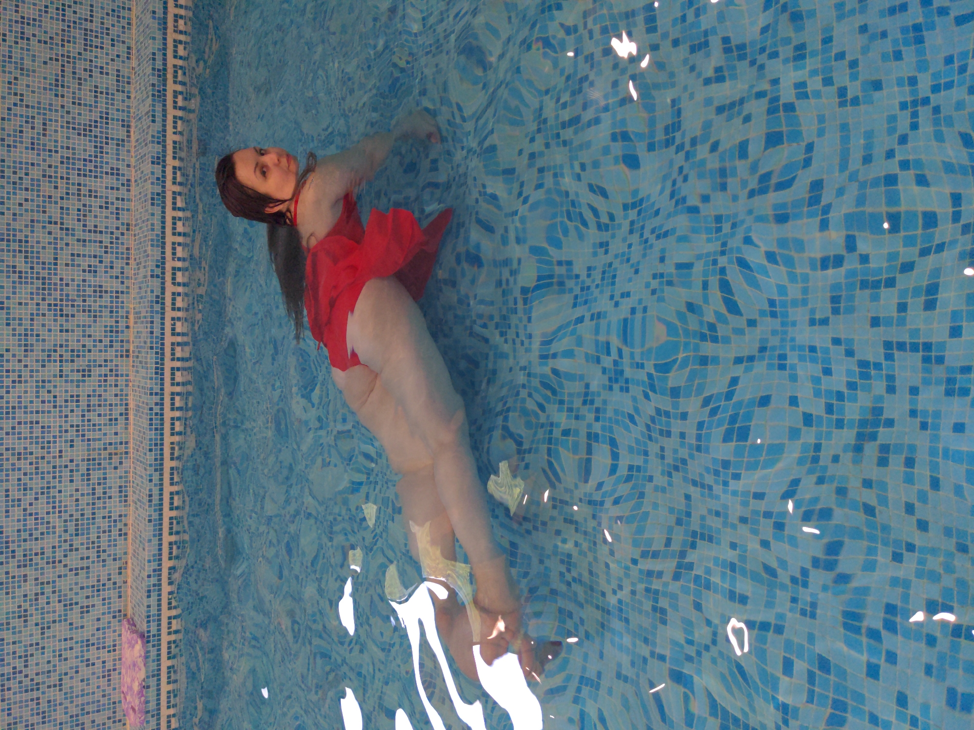 A woman wearing a red dress is swimming in a pool. The pool is tiled with blue and white mosaic tiles. The water is clear and reflects the light. The woman is partially submerged in the water, with her head and shoulders visible above the surface. She is looking towards the camera. The background is a tiled wall. The scene is located in a swimming pool in Holubyne, Ukraine.