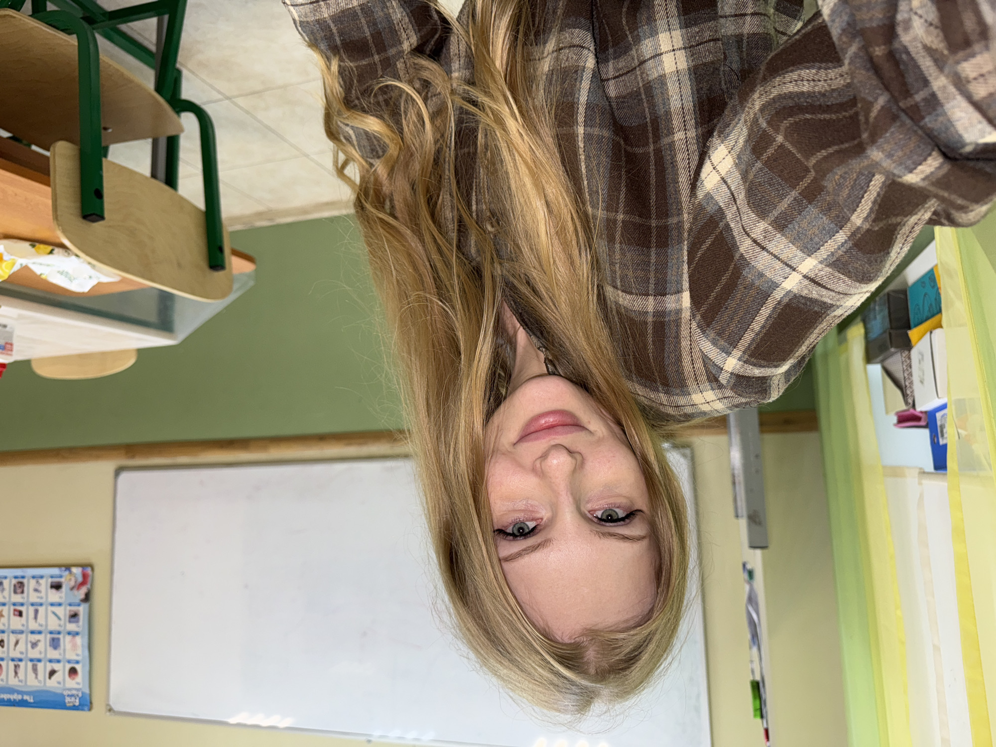The image shows a young woman with long blonde hair, wearing a brown and beige plaid shirt. She is looking directly at the camera with her head tilted. Her eyes are green and she has light makeup on, including eyeliner and mascara. Her lips are slightly parted.
The setting appears to be a classroom. In the background, a whiteboard is visible, along with an alphabet chart on the wall to its left. There is also a desk with green metal legs and wooden surfaces, and a green wall in the background. Some shelves with items on them are visible to the right, with light yellow and green curtains. The lighting suggests it is daytime. There is no discernible weather information. The location is specified as Perm, Russia.