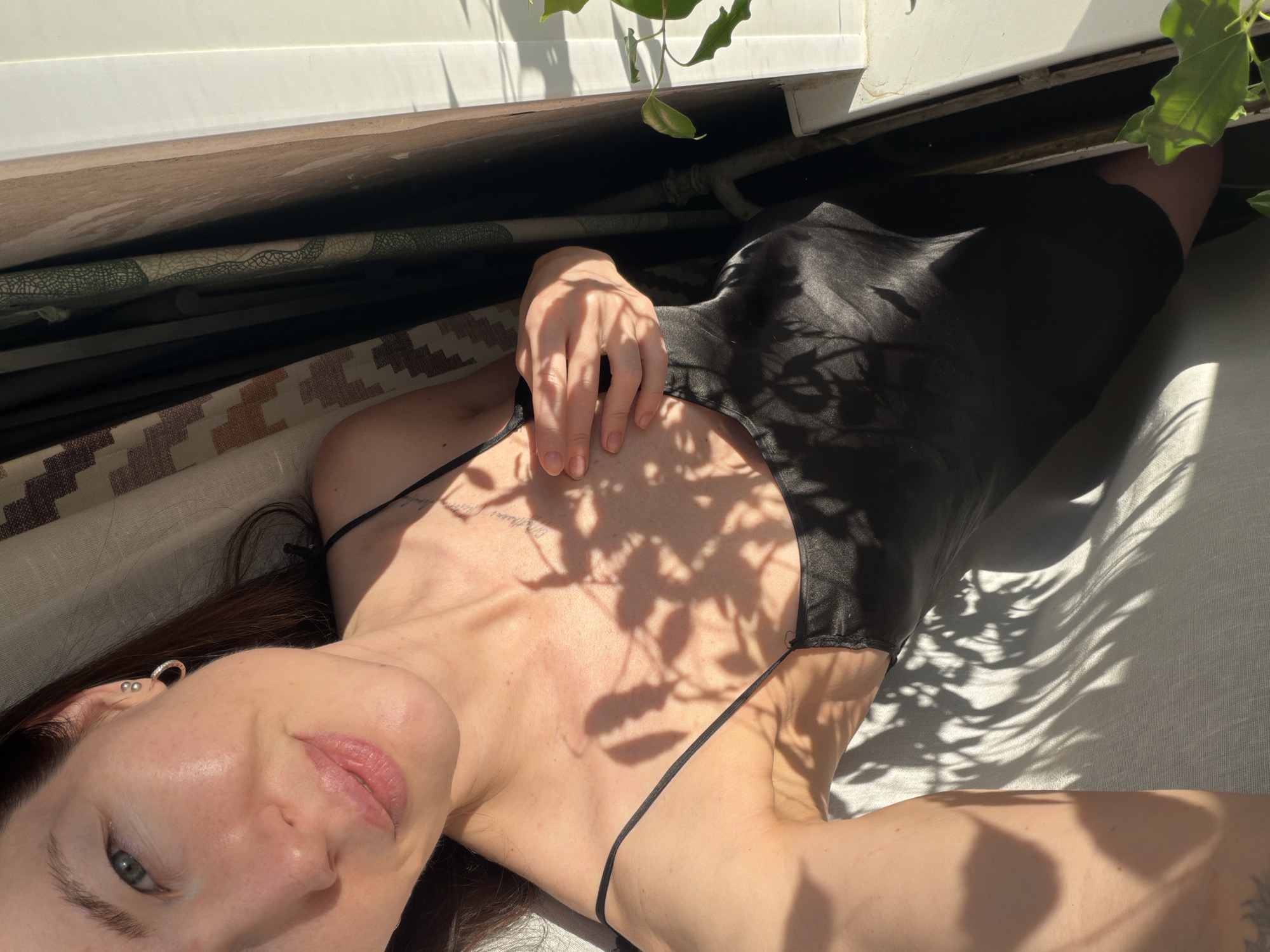A woman is lying down in a room with sunlight filtering through plant foliage, casting shadows on her body and surroundings. She is wearing a black slip dress with thin straps. Her left hand rests on her chest. Tattoos are visible on her upper chest and collarbone area. She has dark hair and light skin. The background includes a window and curtains or blinds with a patterned fabric. The photograph was taken in a room in Perm, Russia.
