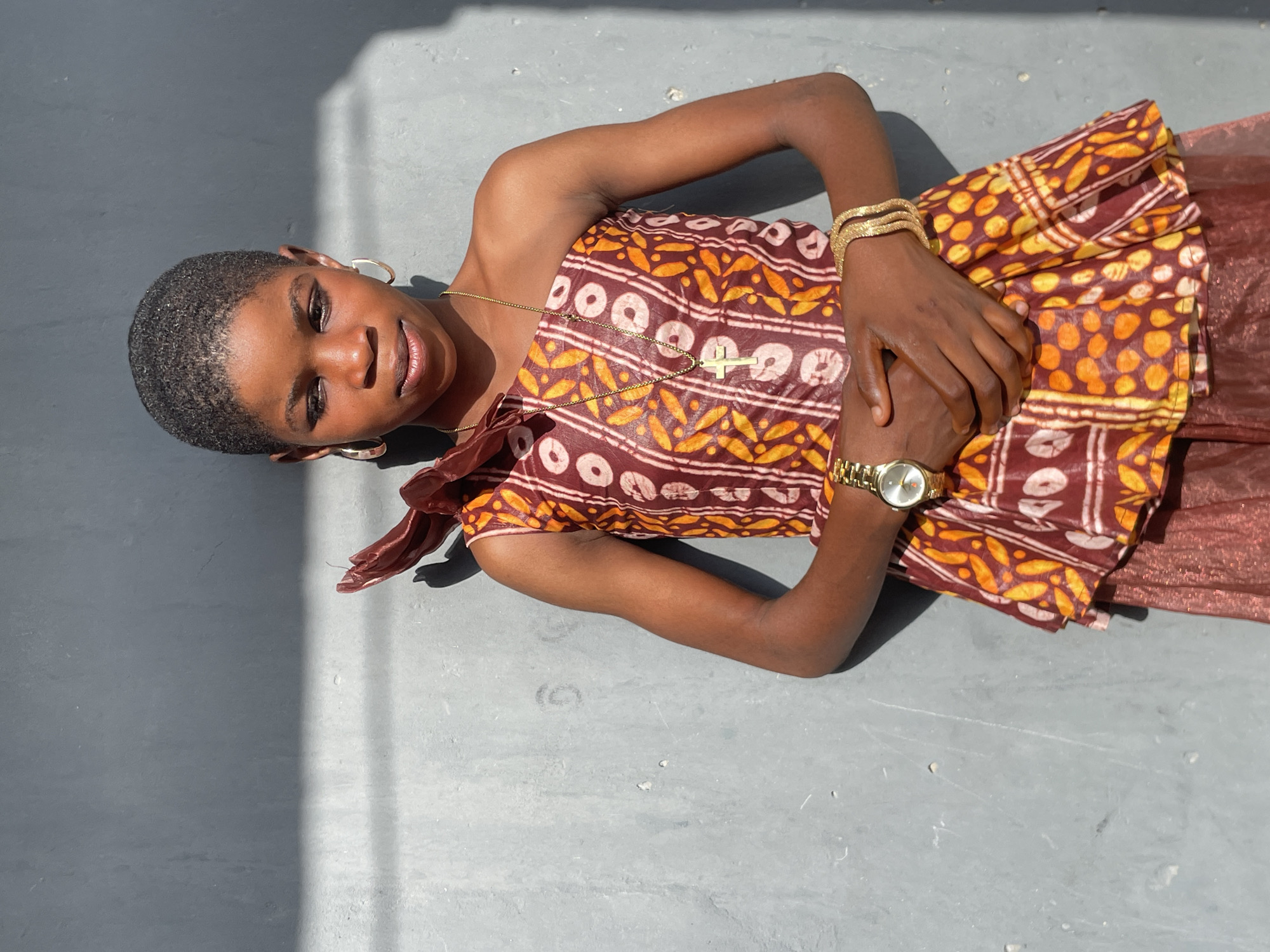 A young person is depicted lying on a light grey, possibly concrete, surface under bright natural light. The image is oriented with the subject's head to the left, looking directly at the camera with a gentle, relaxed smile. They have very short, dark hair, dark skin, and wear light eye makeup and subtle lip gloss.
The subject is dressed in a vibrant, patterned garment. The top portion of the dress features a reddish-brown base fabric adorned with bold yellow and orange patterns, including circular and leaf-like shapes. The lower part of the outfit is a shimmery reddish-brown material.
Accessories include gold-colored hoop earrings, a thin gold chain necklace with a cross pendant, multiple gold-colored bangles on the left wrist, and a gold-toned watch with a round white dial on the right wrist. Their hands are gently clasped over the patterned dress.
The outdoor setting is illuminated by strong, directional sunlight, casting distinct, elongated shadows across the grey ground, particularly on the upper left and lower right areas of the frame. This indicates a clear, sunny day, likely during midday or early afternoon. The overall scene conveys a calm and pleasant atmosphere. No visible text is present in the image. The location context is Badagry, Nigeria.