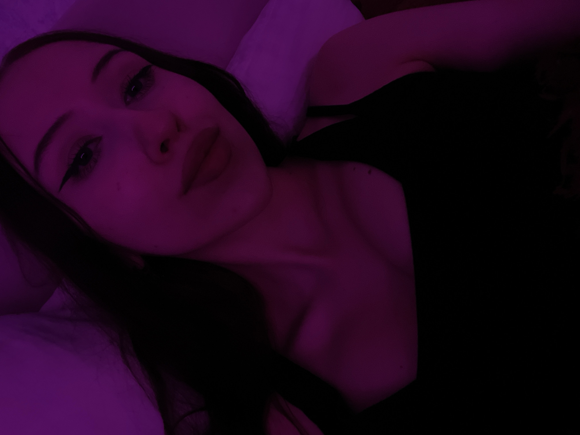 The image is a close-up selfie of a young woman with dark hair, wearing a black tank top. She has eyeliner and lipstick on. The lighting is predominantly purple, casting a dramatic mood over the scene. The background appears to be a bed with a white pillow. There are no discernible elements that would indicate a specific location in Votkinsk, Russia. The focus is solely on the woman's face and upper body.