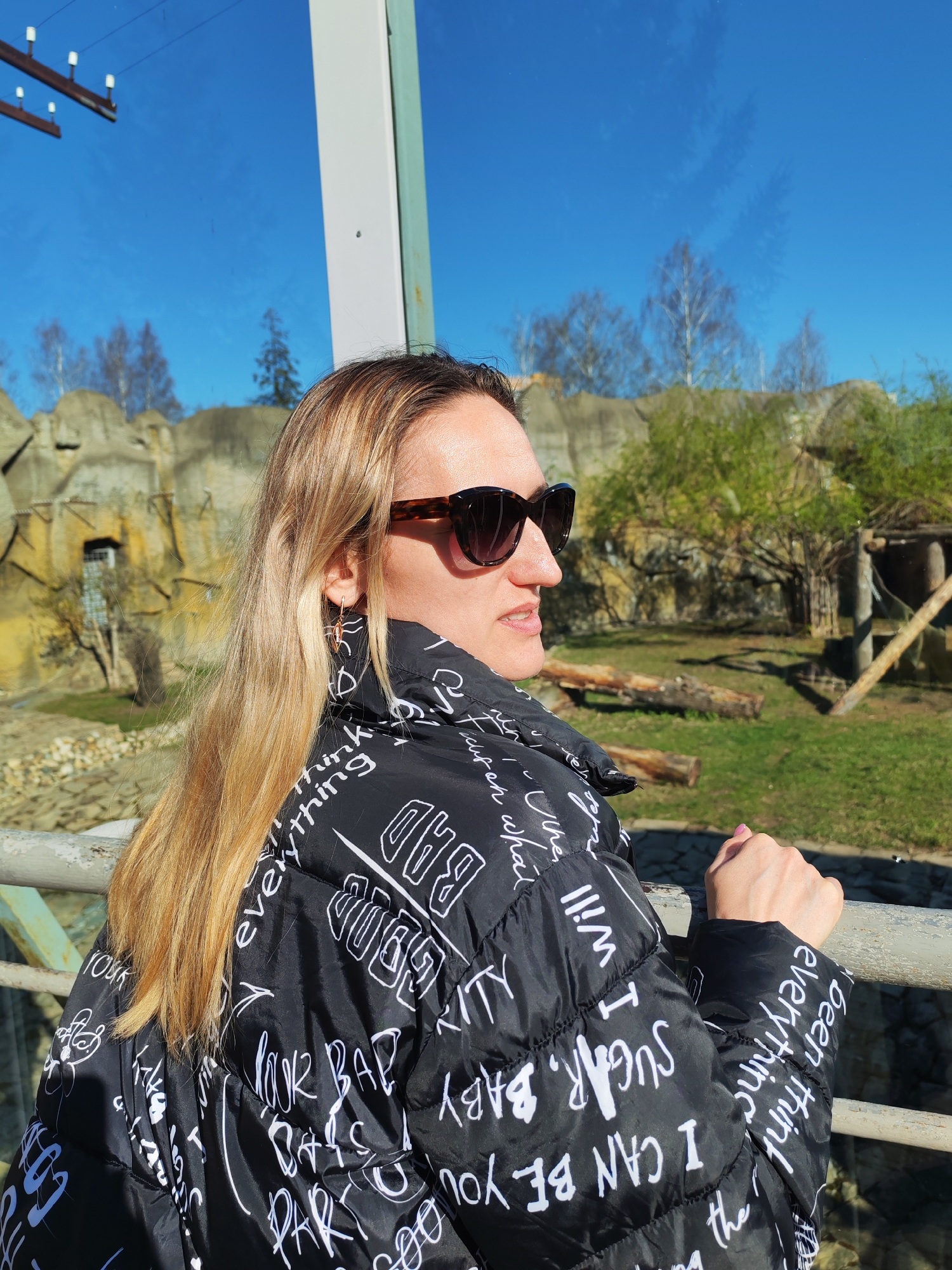 The image shows a woman at the Izhevsk Zoo in Russia. She is standing indoors, near a window, looking out at the outdoor enclosure. The woman is wearing sunglasses and a black jacket with white lettering. The enclosure has a rocky background and contains logs, trees, and a wooden shelter. The sky is blue and visible through the trees. The scene is well-lit.
