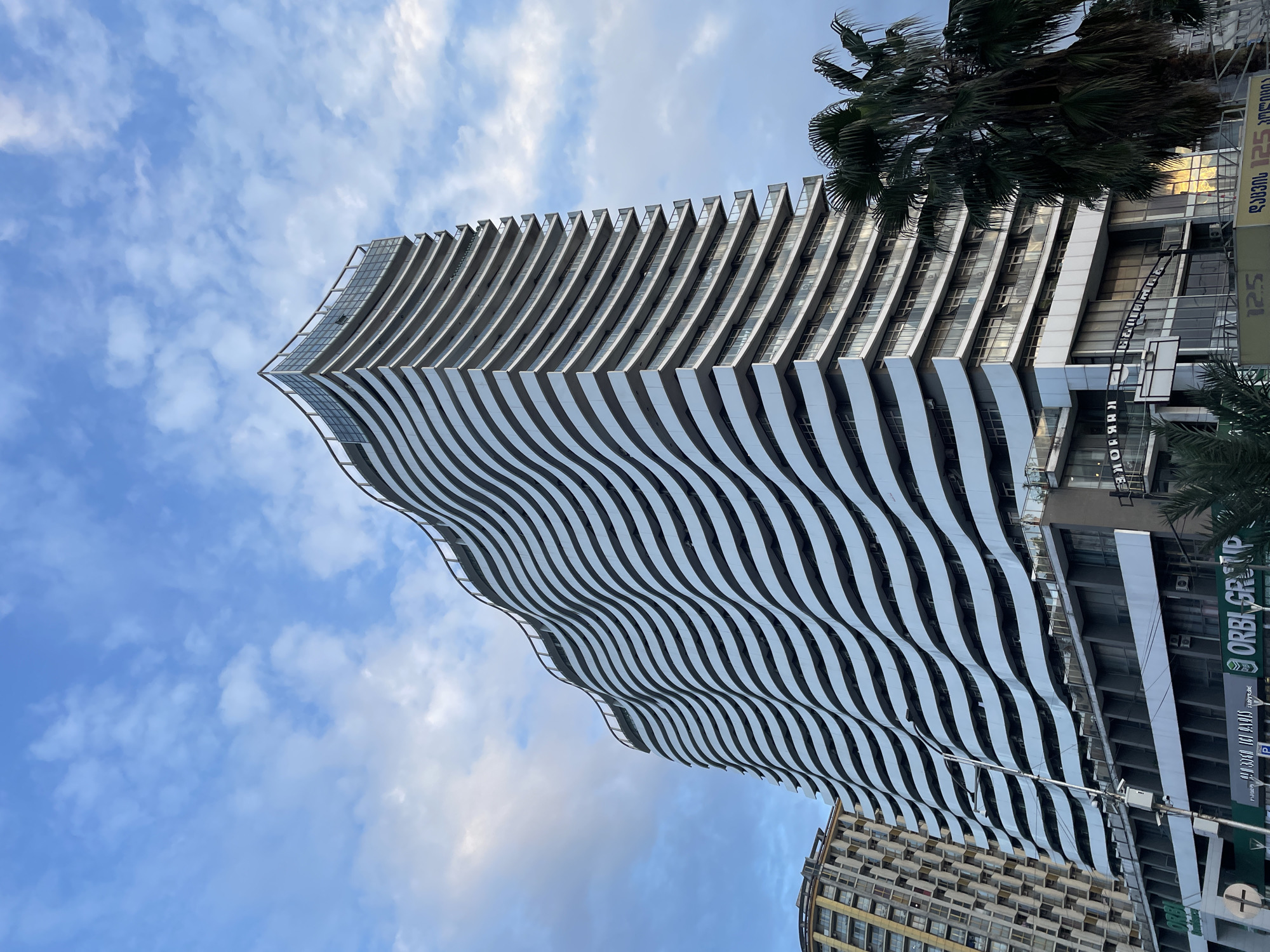 The photograph shows a tall, modern building in Batumi, Georgia, characterized by its wave-like facade of white horizontal stripes. The building is angled upward against a backdrop of a partly cloudy sky. A palm tree is present to the right of the building. Adjacent buildings and street signs are visible on the right and lower portion of the image, providing context to the urban environment.