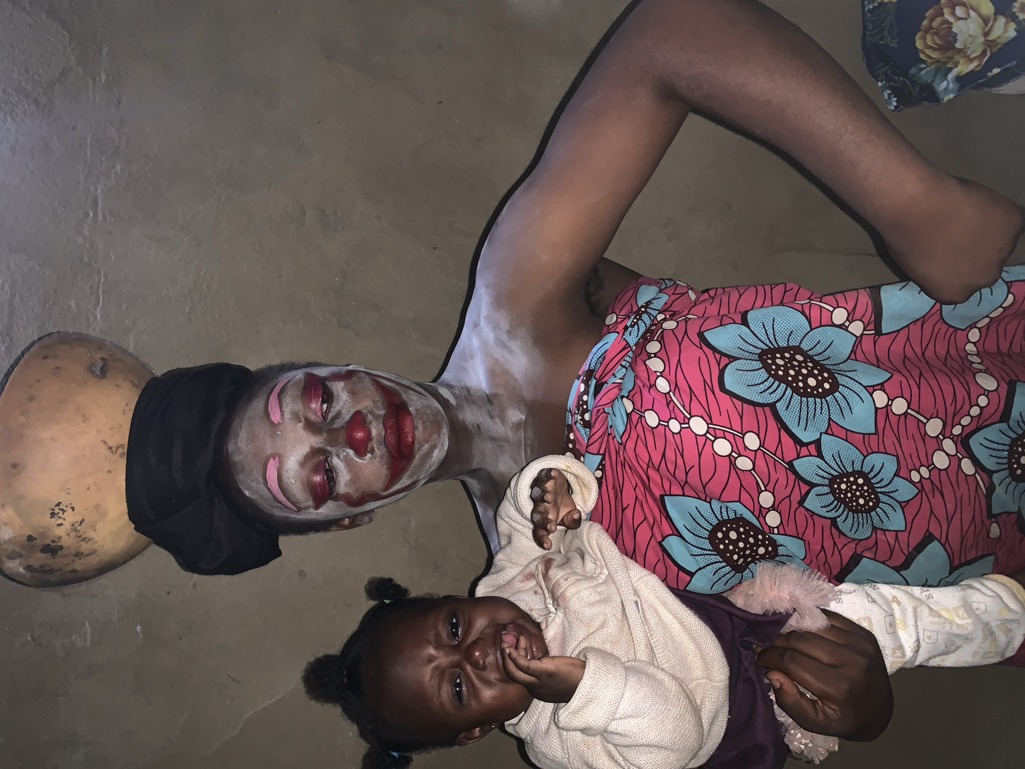 In Agban, Nigeria, a woman with clown-like makeup, including white powder and red accents on her eyes and lips, balances a gourd on her head. She is wrapped in a vibrant pink and blue floral fabric. She holds a distressed baby, who is crying and has its hand in its mouth. The baby is wearing a cream-colored sweater. The background is a plain, textured wall.