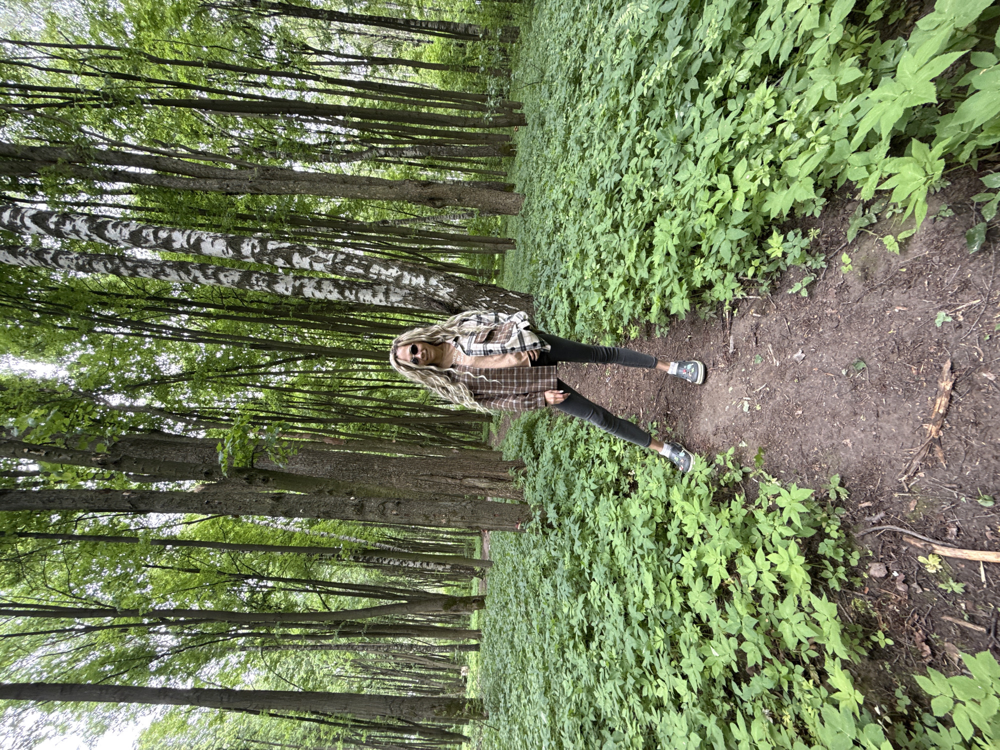 A woman with long blonde hair and sunglasses stands in the middle of a dirt path in a forest. She is wearing a plaid shirt, black pants, and sneakers. The forest is dense with trees and green foliage. The location is Rusanovo, Russia.