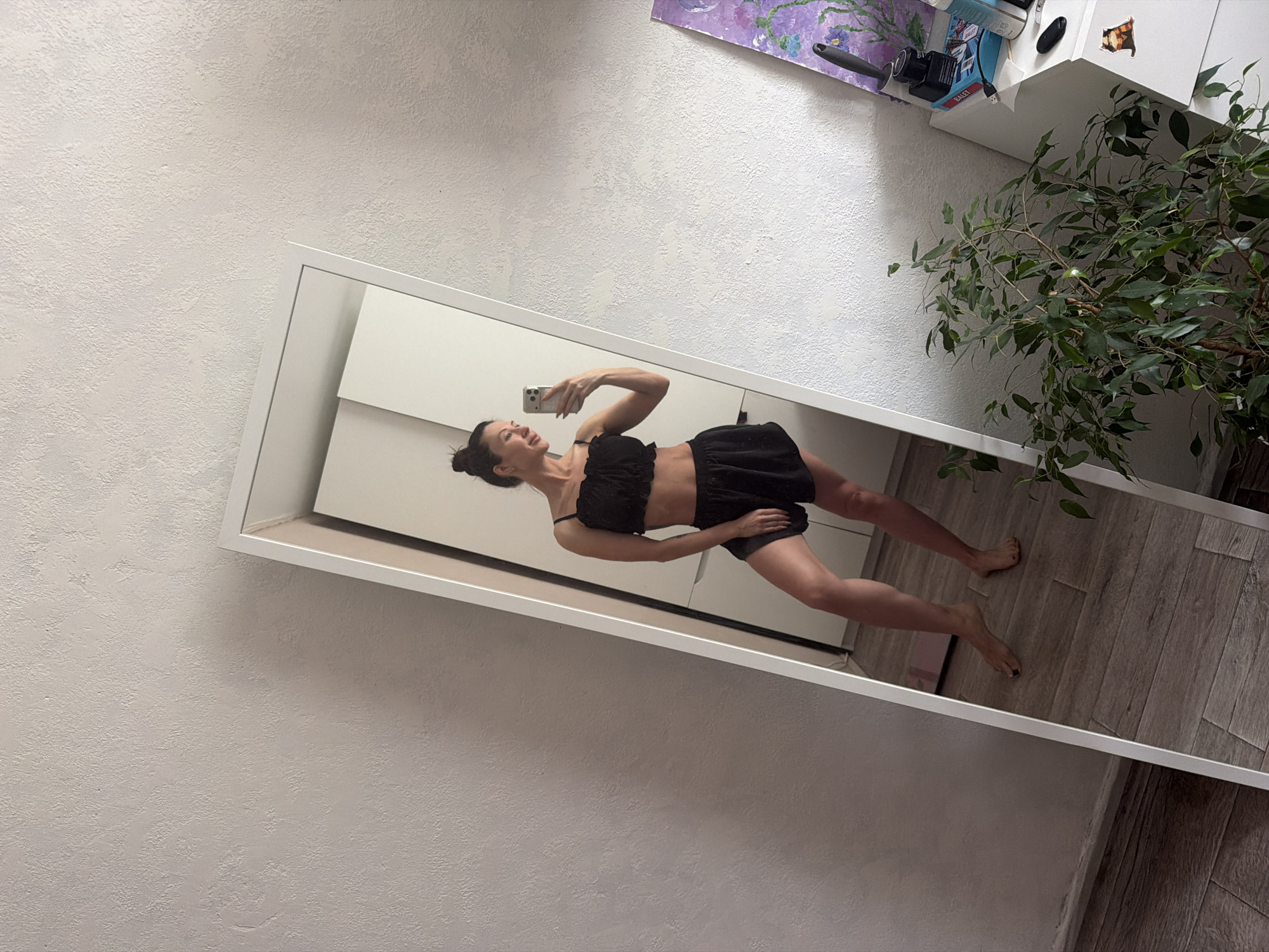 The image captures a full-body mirror selfie taken indoors, likely in Perm, Russia. A young woman with dark hair tied in a bun is reflected in a large, white-framed mirror, posing with a smartphone in her right hand to take the picture. She is wearing a black two-piece outfit, consisting of a ruffled crop top and matching shorts, highlighting her toned physique. Her head is tilted back slightly, and she has a happy, confident expression with an open-mouthed smile.
The scene depicts a contemporary room with a textured, light-colored wall visible both directly and in reflection. The floor in the reflection is covered with wooden-patterned laminate or planks. Above the woman's reflection, a white shelf or cabinet is visible, holding various items. These include a vibrant purple and green abstract painting or artwork, a black camera or electronic device, a small blue package partially showing the text "GALET," a white power adapter with "USB" visible, a black computer mouse or similar device, and a potted plant with lush green leaves (possibly a Ficus). The overall impression is a personal and casual moment captured in a modern home setting. The time of day appears to be bright, suggesting daytime.