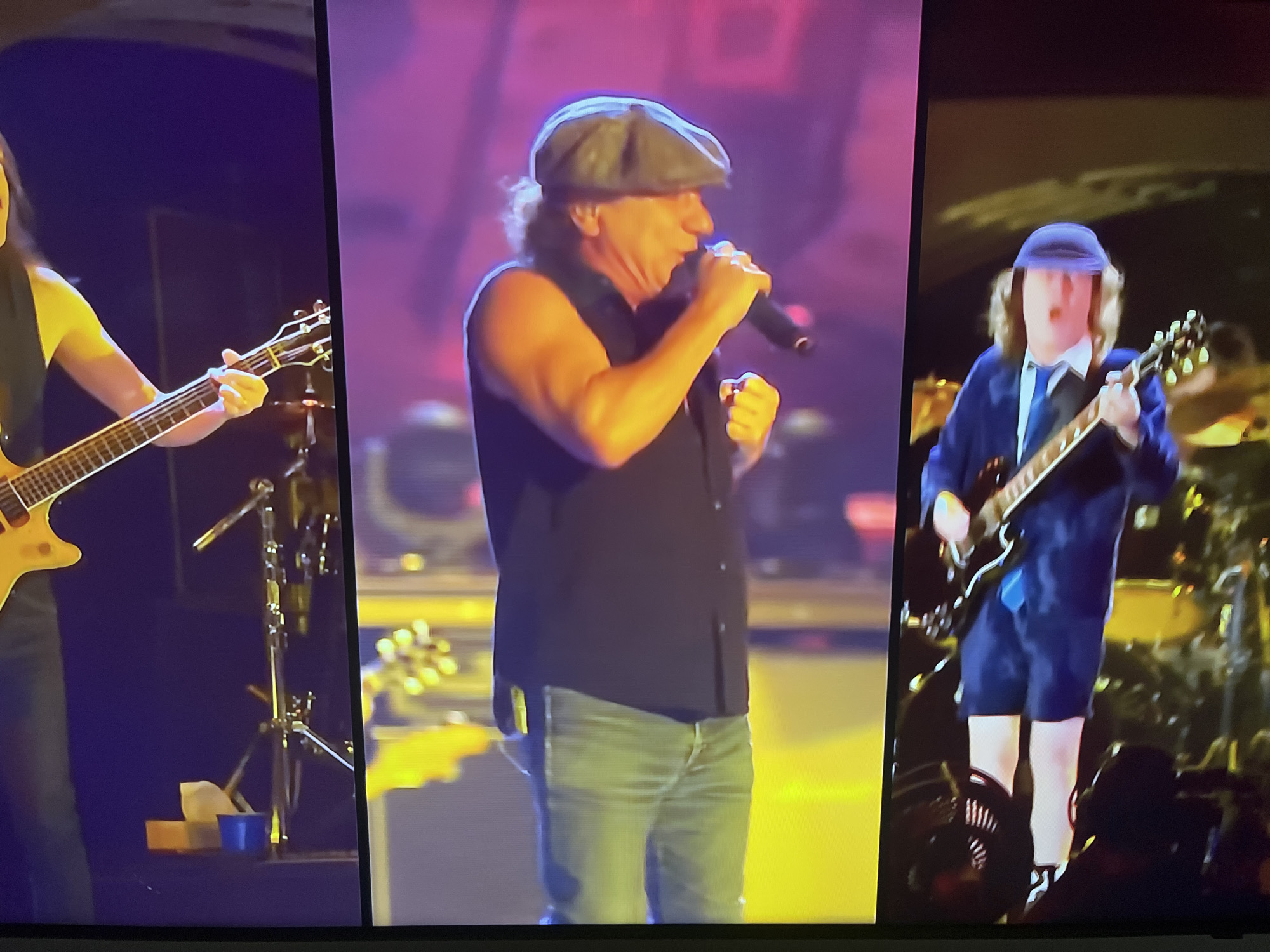 The image shows a split-screen view of a live music performance. On the left, a person is playing a yellow electric guitar. In the center, a person wearing a black sleeveless shirt, blue jeans, and a flat cap is singing into a microphone, holding one fist up. On the right, a person in a school uniform, consisting of a blue jacket, shorts, white shirt, and blue tie, is playing a black electric guitar. All three individuals appear to be on a brightly lit stage with purple and yellow lighting. There is no information in the image to suggest this location is in Kharkiv, Ukraine.