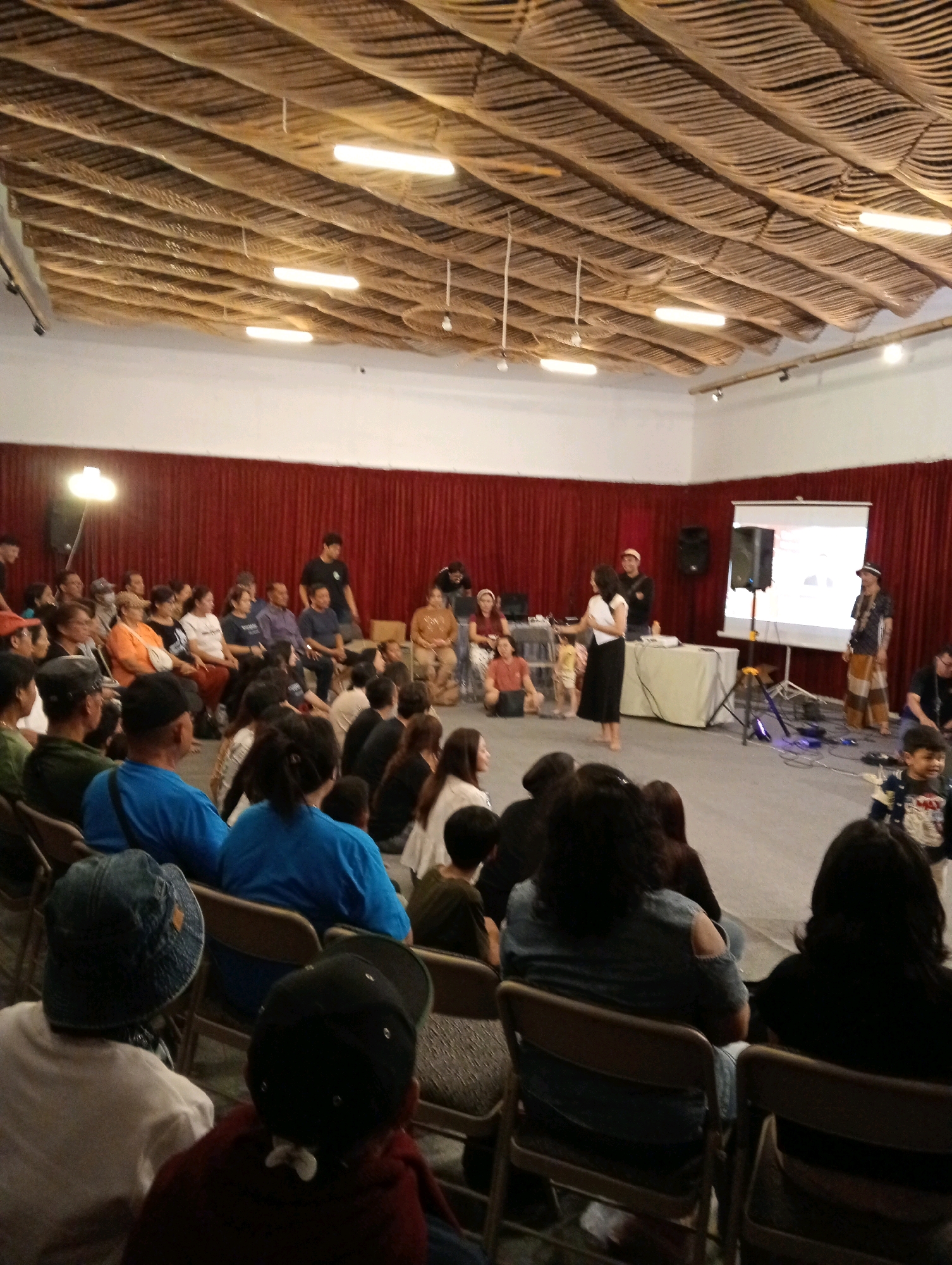 The image depicts an indoor event with a seated audience facing a stage area. The ceiling is constructed with a woven material, possibly palm fronds, with linear lighting fixtures. A red curtain forms the backdrop of the stage. In the foreground, several audience members are visible, mostly from behind, seated in rows of chairs. The audience members are of various ages and genders, some wearing casual clothing and hats. On the stage, there are people standing and interacting. A projector screen is set up, displaying an image. Two speakers are positioned near the screen. There's a table covered with a cloth, and various electronic equipment. The overall atmosphere suggests a presentation, performance, or gathering. The location is identified as Pacet, Indonesia, though no specific landmarks confirm this within the image itself.