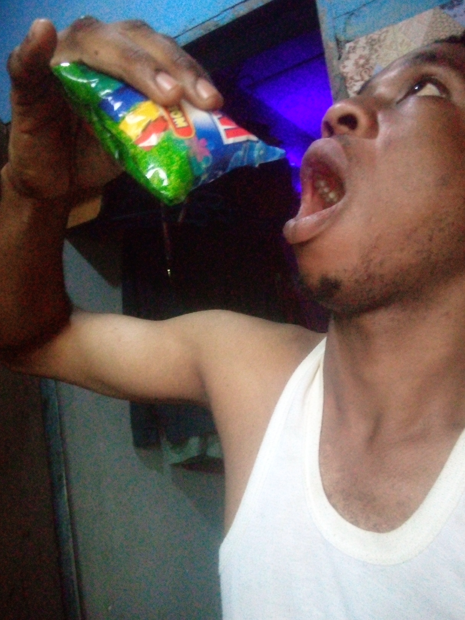 A person in a white tank top is drinking from a small, green and blue tetra pak-like package. The person is positioned at an angle, with their head tilted back and mouth open, as they drink. The background is blurry and dimly lit. The location and city are unspecified.