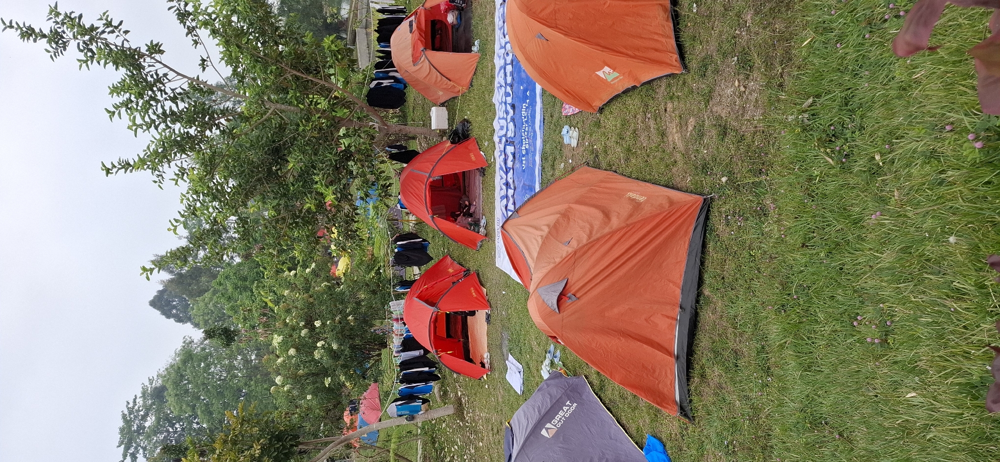 The image depicts a campsite with multiple tents set up on a grassy area, likely a park or open field, under a cloudy sky. In the foreground, several tents are visible, including a prominent orange tent and a gray tent with "GREAT OUT DOOR" branding. In the middle ground, two red tents are open, revealing sleeping mats inside, and a clothesline strung between trees is laden with laundry. Further in the background, more tents of various colors, including red and yellow, are scattered across the landscape. Trees and foliage surround the campsite, and a large blue banner with Indonesian text is partially visible on the ground in the foreground. The overall scene suggests a camping event or gathering.