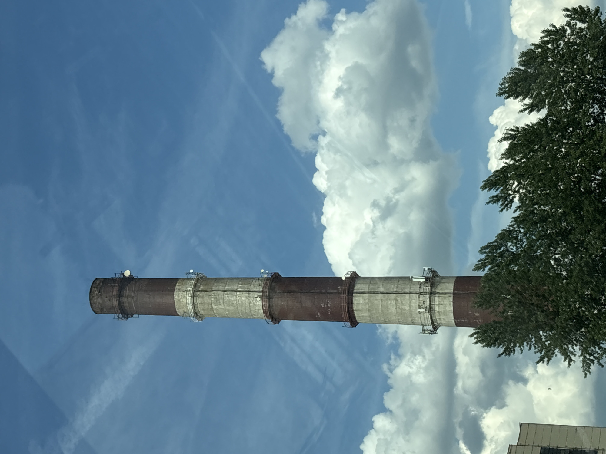 A tall, cylindrical industrial chimney, likely part of a combined heat and power plant (CHP), dominates the scene under a bright blue sky with scattered cumulus clouds and visible aircraft contrails. The chimney exhibits alternating horizontal bands of weathered, light-gray concrete and reddish-brown discoloration or paint, suggesting a pattern of five segments: a dark top, followed by a light, dark, light, and dark segment down to the visible base. Multiple metal platforms with railings encircle the chimney at various heights, each hosting several white, dish-like and cylindrical telecommunication antennas. The top edge of the chimney appears dark and slightly flared. In the lower left quadrant, a portion of a utilitarian, light-colored building with a dark rectangular recess is partially visible. The lower section of the image features the dense green foliage of several deciduous trees, partially obscuring the lower part of the chimney. No individuals or discernible activities are depicted. The scene is illuminated by natural daylight. This structure is located in Saint Petersburg, Russia, specifically identifiable as a chimney belonging to an industrial heating plant, potentially Polyustrovskaya CHP (CHP-14) in the Vyborgsky District, known for its distinct appearance and repurposed use for communication infrastructure.