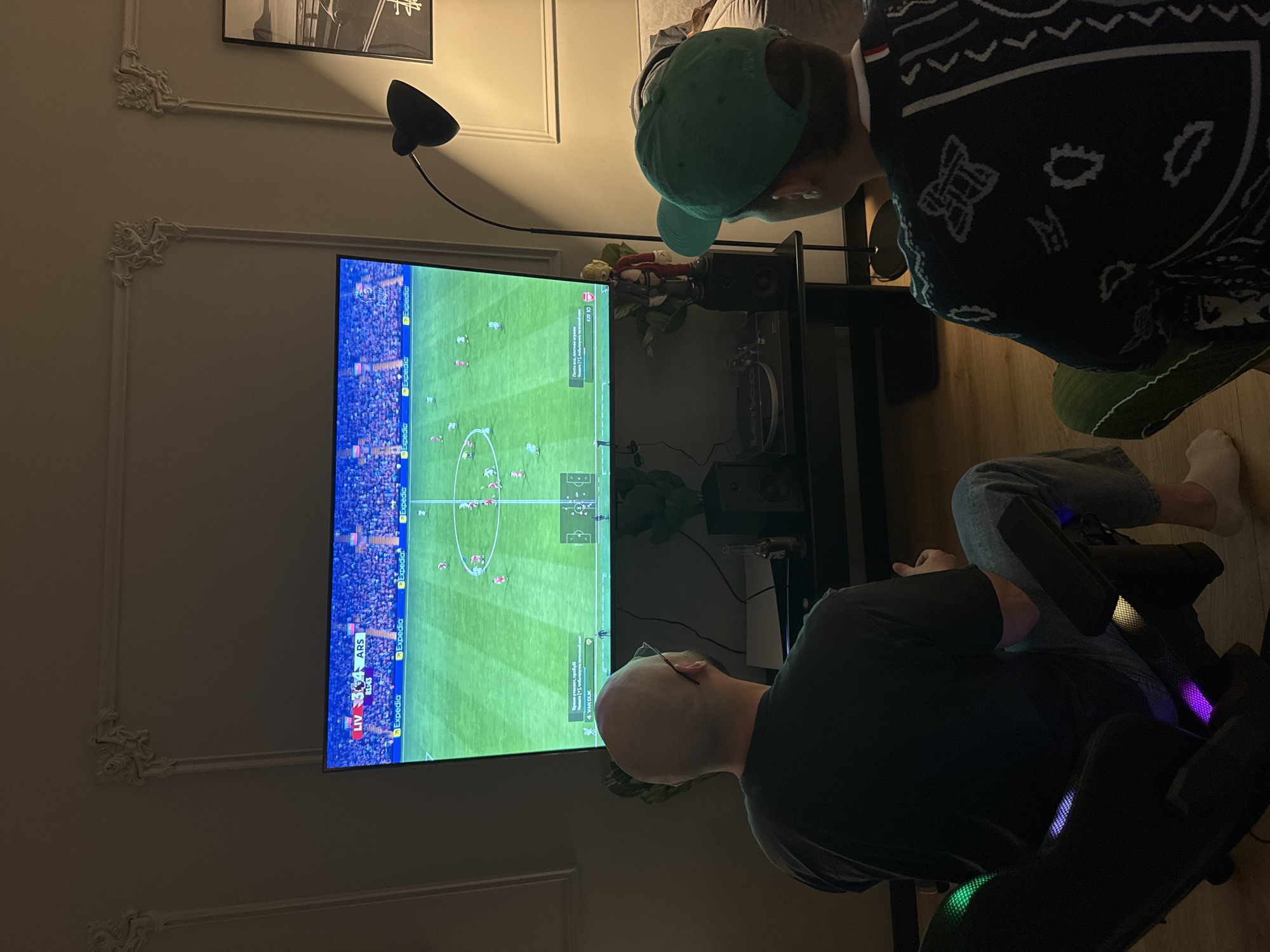 Two people are watching a soccer game on a large television. One person is wearing a green baseball cap and a patterned sweater, while the other is seated in a gaming chair. The TV is displaying a soccer match with a score of Liverpool 3, Arsenal 4. A lamp is positioned above the TV. The scene is located in an indoor room with decorative molding on the walls. The city is not shown in the image.