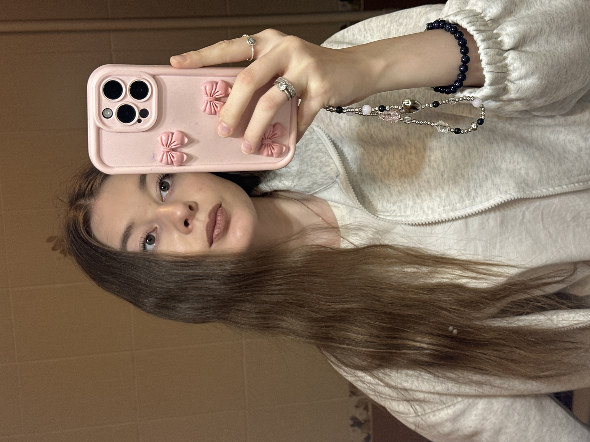 A young woman is taking a selfie. She is holding a pink phone case decorated with bows. She is wearing rings on her fingers and a bracelet on her wrist. She has long, brown hair. The background appears to be a tiled wall, typical of a bathroom. There is no information available to suggest that the image was taken in Kazan, Russia.