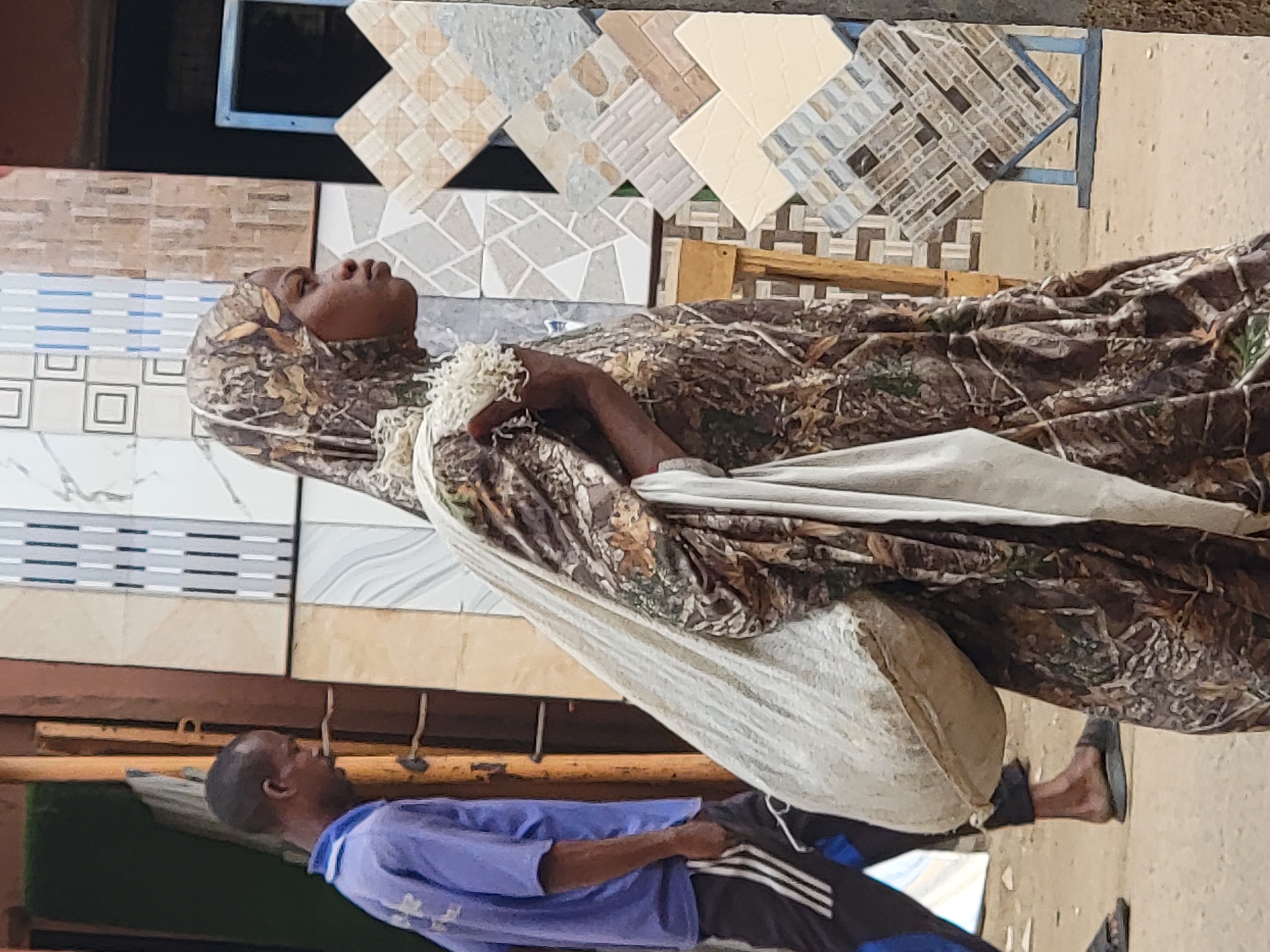 In the city of Maiburiji, Nigeria, the scene captures a person, presumably female, wearing a camouflage headscarf and garment, lying in a hammock-like contraption. This makeshift bed is suspended over a man clad in a blue shirt and athletic pants with white stripes, lying on what appears to be a padded surface. The backdrop consists of tile samples mounted on a display.