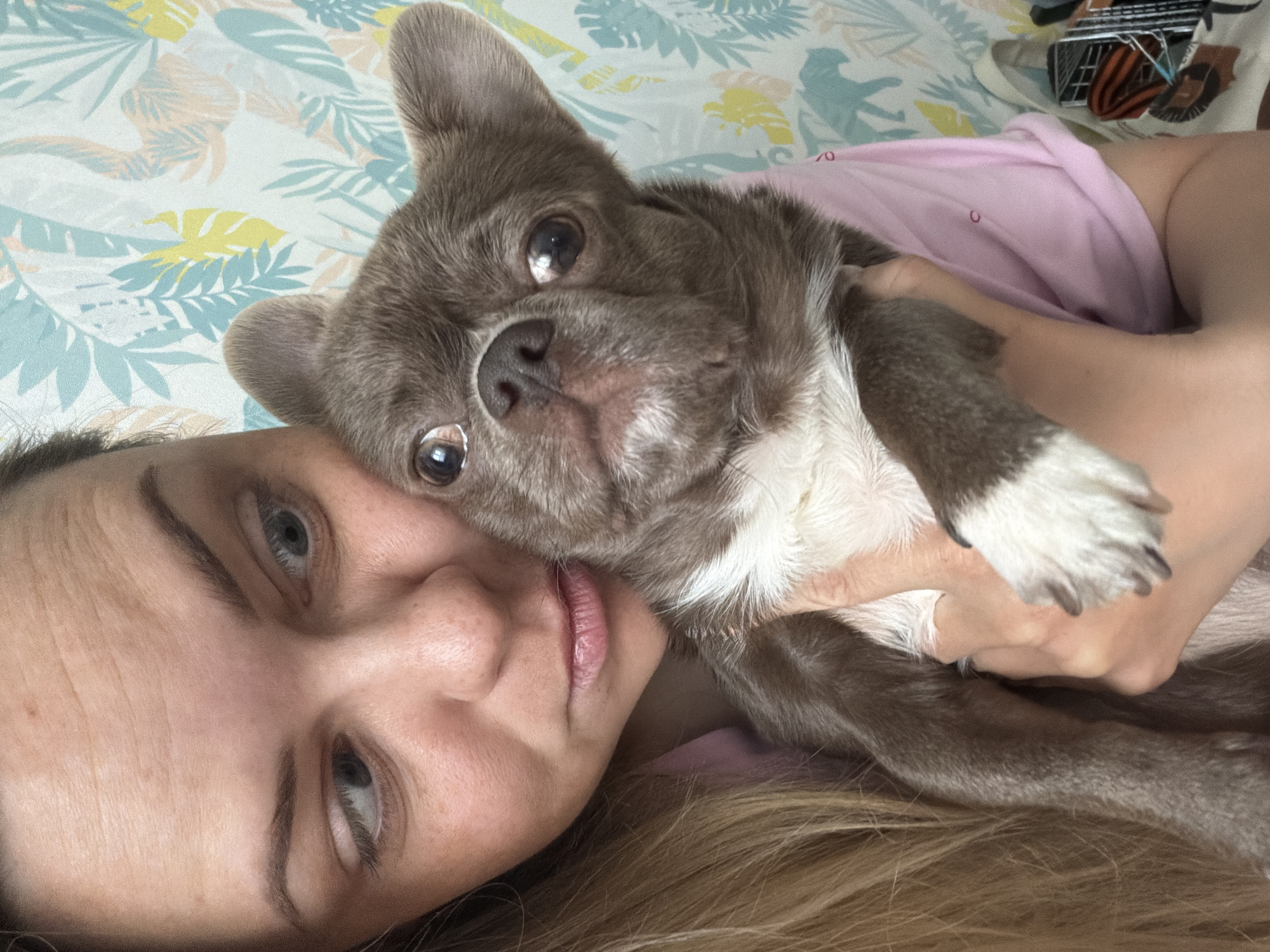 A woman with blonde hair and light eyes lies on a bed with a floral-patterned sheet, holding a gray and white dog in her arms. The dog is looking directly at the camera, while the woman is looking slightly off to the side. The woman is wearing a pink shirt. The location cannot be determined, as there are no indicators to confirm the city of Votkinsk, Russia.