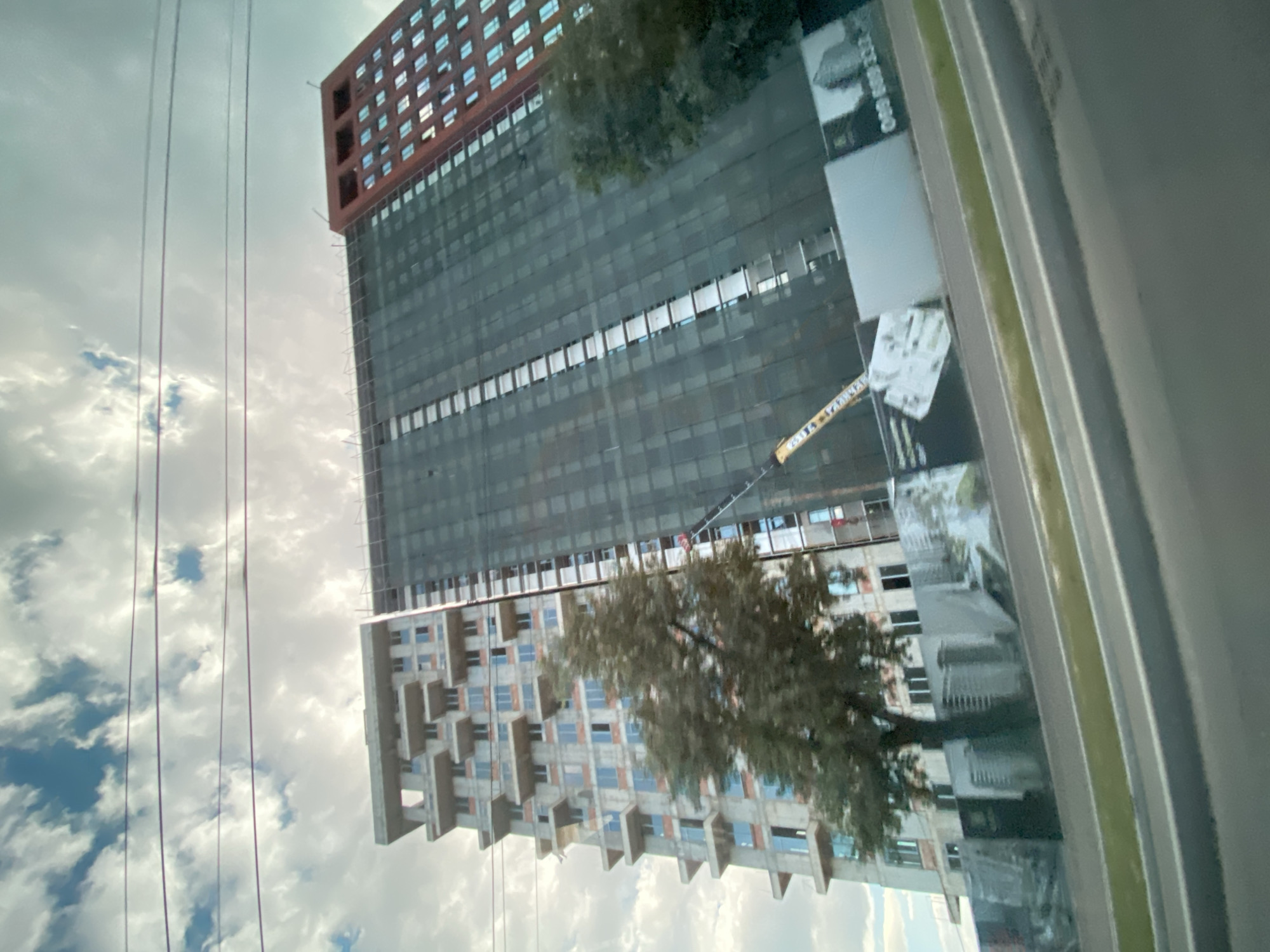 An unfinished, multi-story building is under construction in Chișinău, Moldova. The building on the left is partially clad with concrete and has visible balconies. The larger building to the right is covered with dark netting and appears to be more advanced in its construction. A yellow crane is visible extending from the side of the netting. In the foreground, there is a tree and a construction fence with promotional signage, one of which displays a phone number. The scene is viewed from a low angle, possibly from a moving vehicle on a road, with a slightly tilted perspective. The sky above is cloudy with patches of blue.