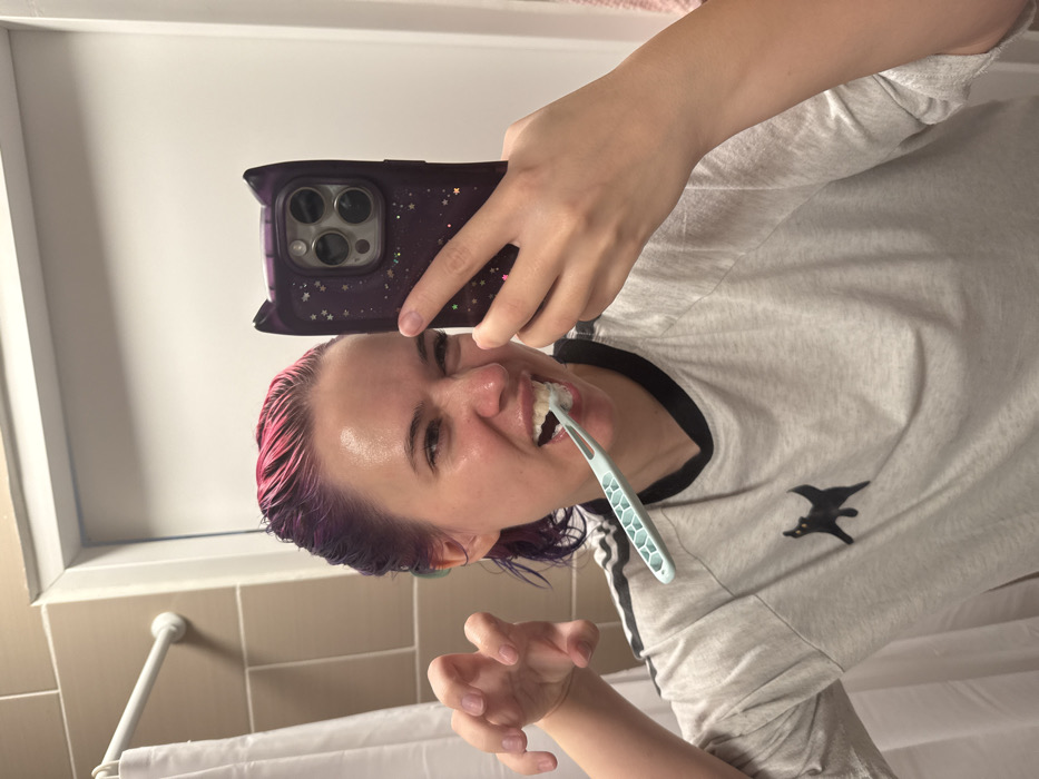 A female is depicted in a bathroom in the City of Zagreb, Croatia, brushing her teeth while taking a mirror selfie with a smartphone. She has wet, multi-colored hair, predominantly pink at the front fading to purple. Her face appears moist and she exhibits a playful grimace, revealing teeth and the light blue/green toothbrush. She wears a light grey short-sleeved t-shirt with black trim on the collar and a black stylized cat emblem on the left chest. Her right hand holds a smartphone, encased in a dark purple, cat-eared case with a glitter effect, displaying a triple-lens camera array. Her left hand is raised in a semi-clenched, claw-like gesture. The background features light beige wall tiles, a white shower curtain on a white rod, and a white door frame.