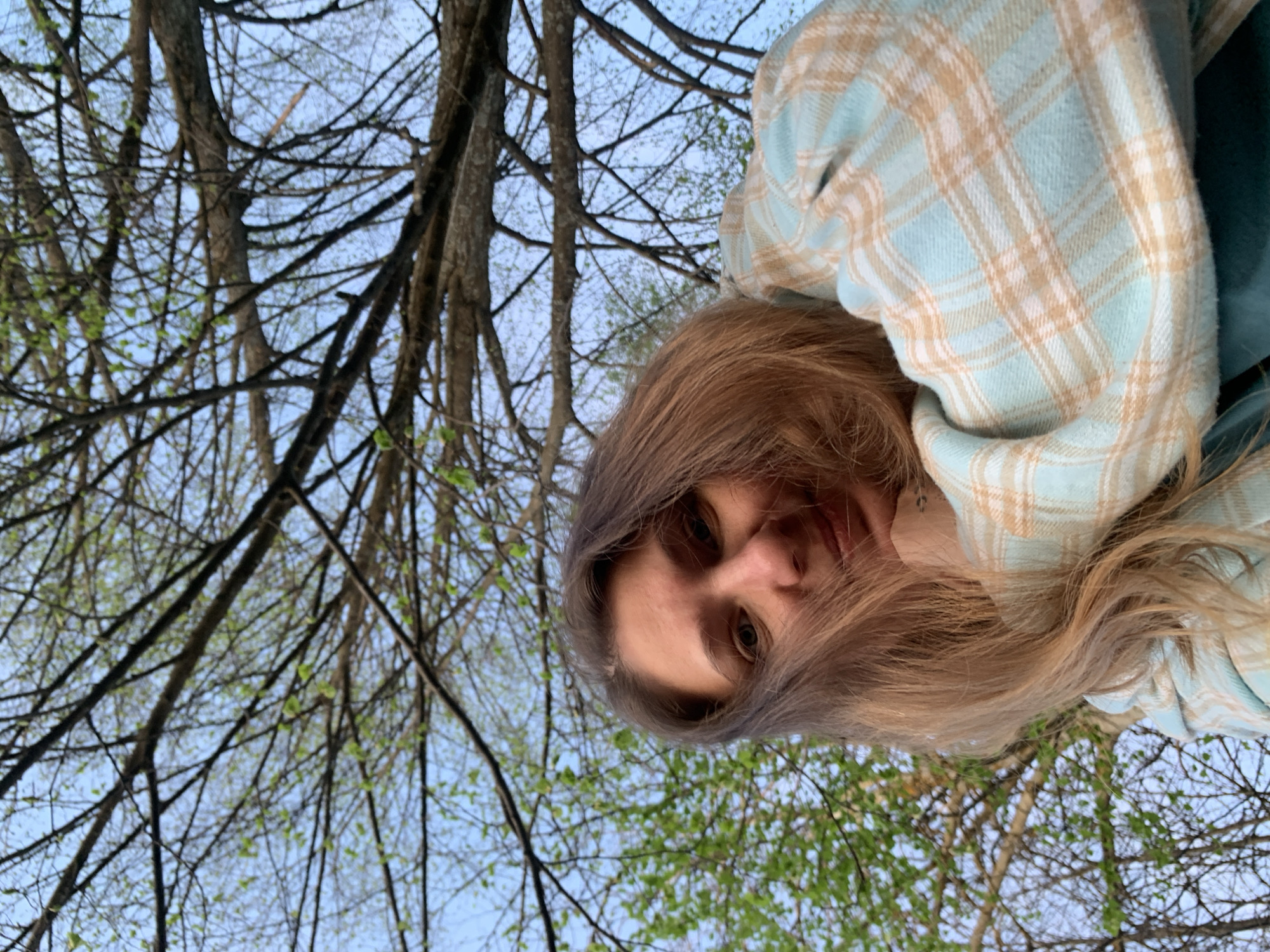 A woman with long, light brown hair wearing a blue and tan plaid shirt is looking down at the camera. Behind her are bare tree branches reaching towards a light blue sky, with some new green leaves visible. The location appears to be an outdoor area, possibly a wooded area or park in Zasechnyi, Russia.
